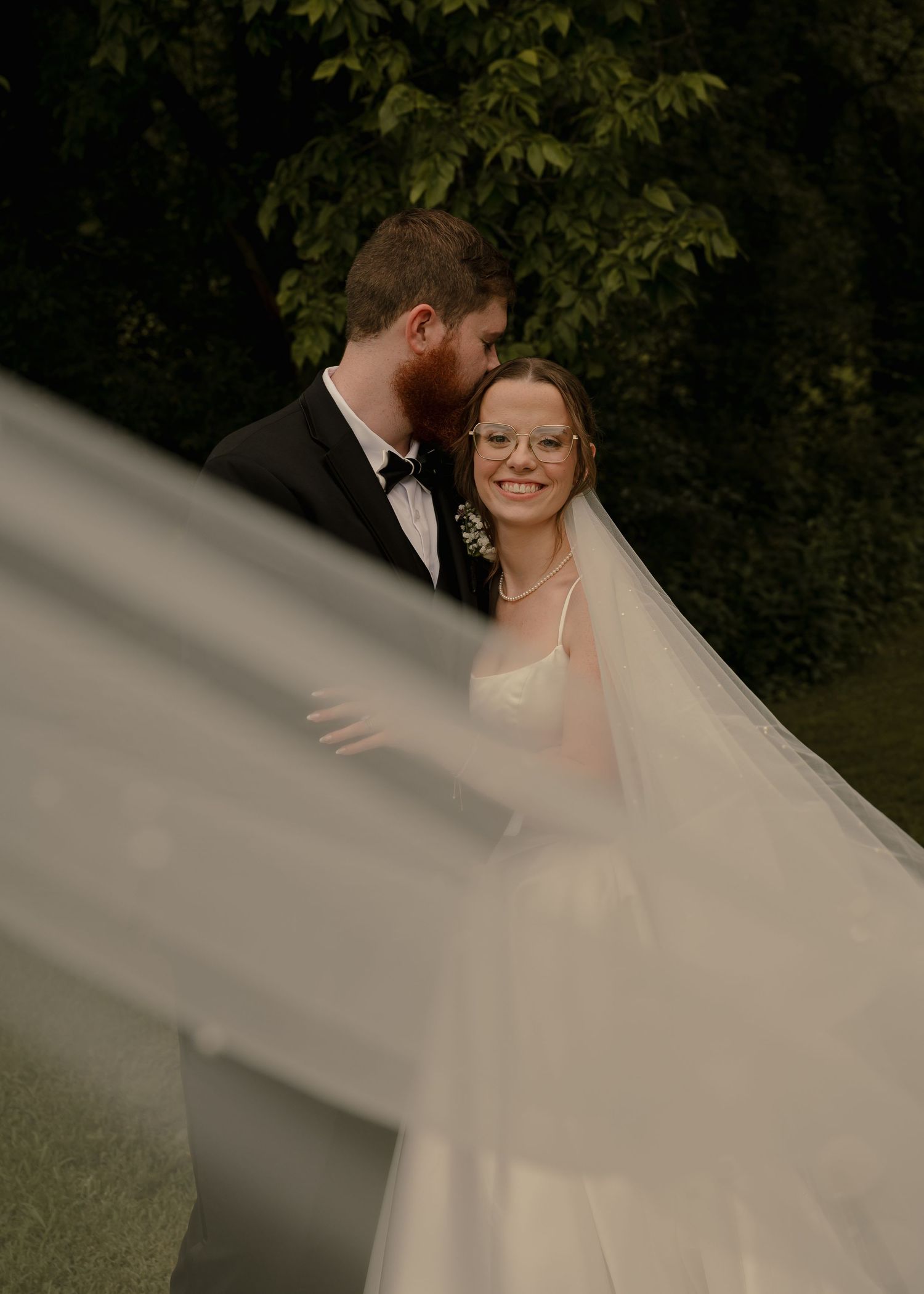 Smiling bride in glasses with groom in black tuxedo framed by flowing veil outdoors.