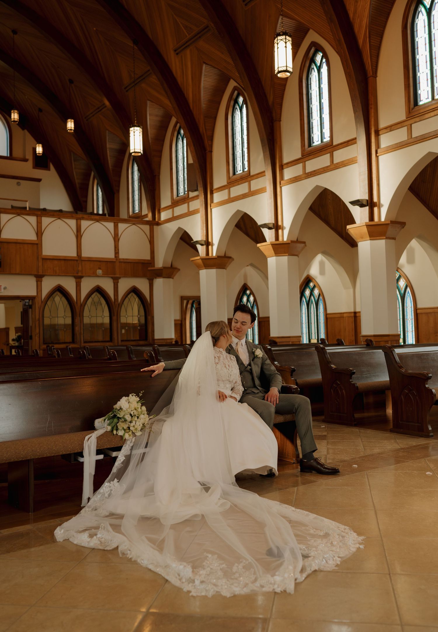 Bride and groom sitting together on church pews beneath soaring Gothic arches with stained glass windows.