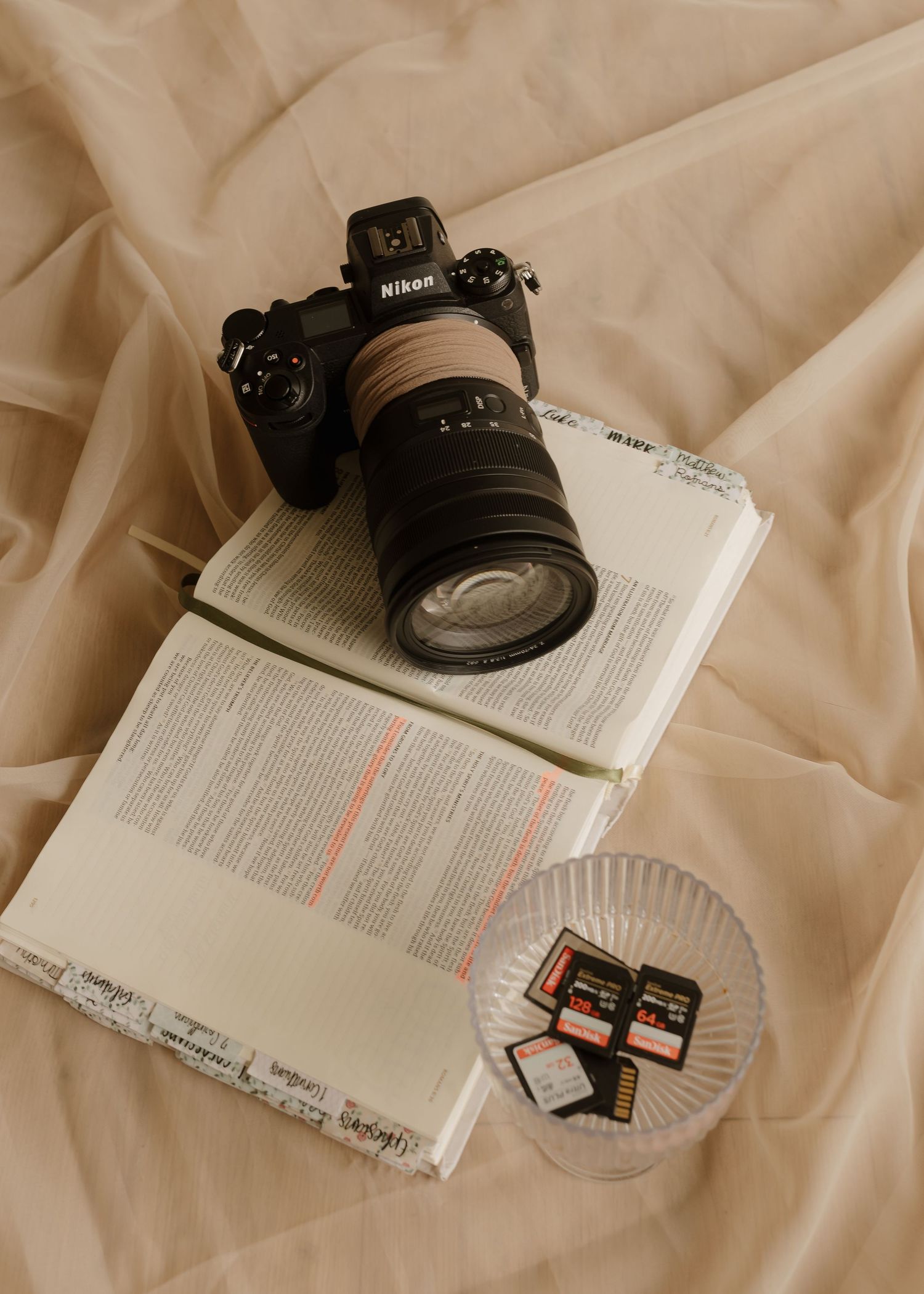 Nikon camera resting on open book beside memory cards on soft beige fabric background.