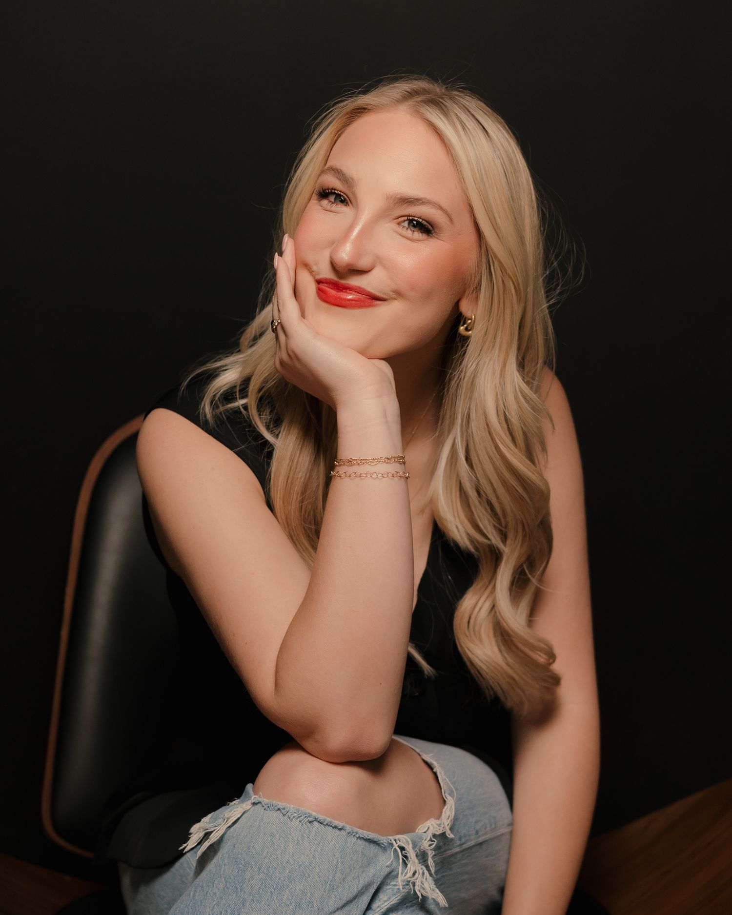 Smiling blonde woman with red lips sitting in leather chair against dark background.