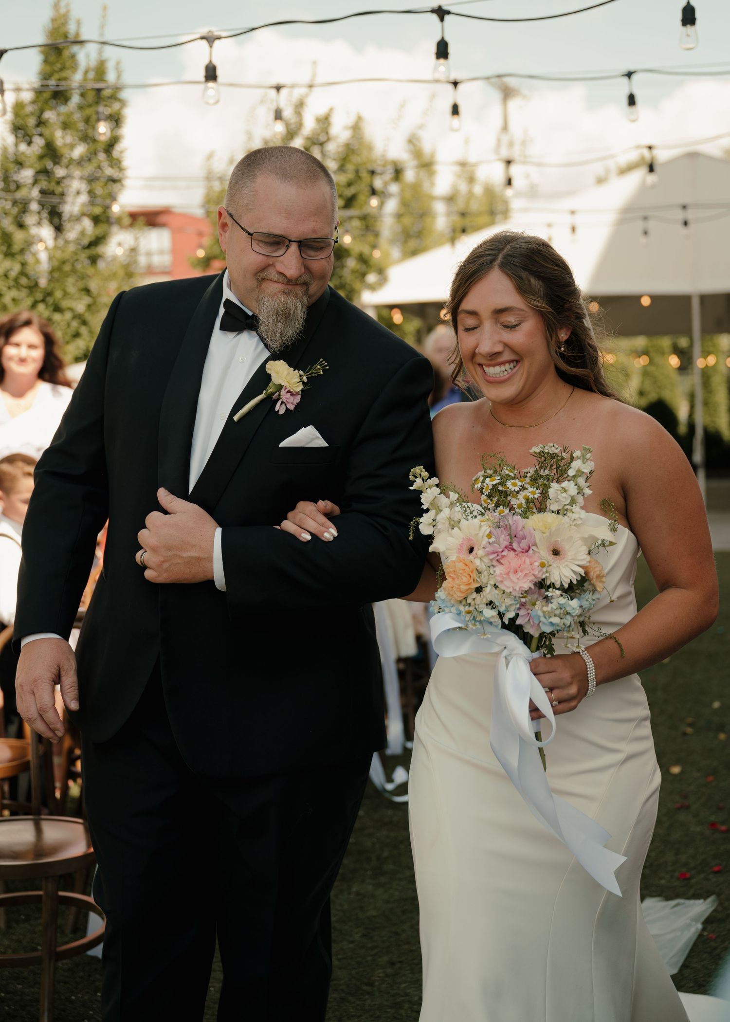 Smiling bride walking down aisle with escort in tuxedo at outdoor evening wedding ceremony.