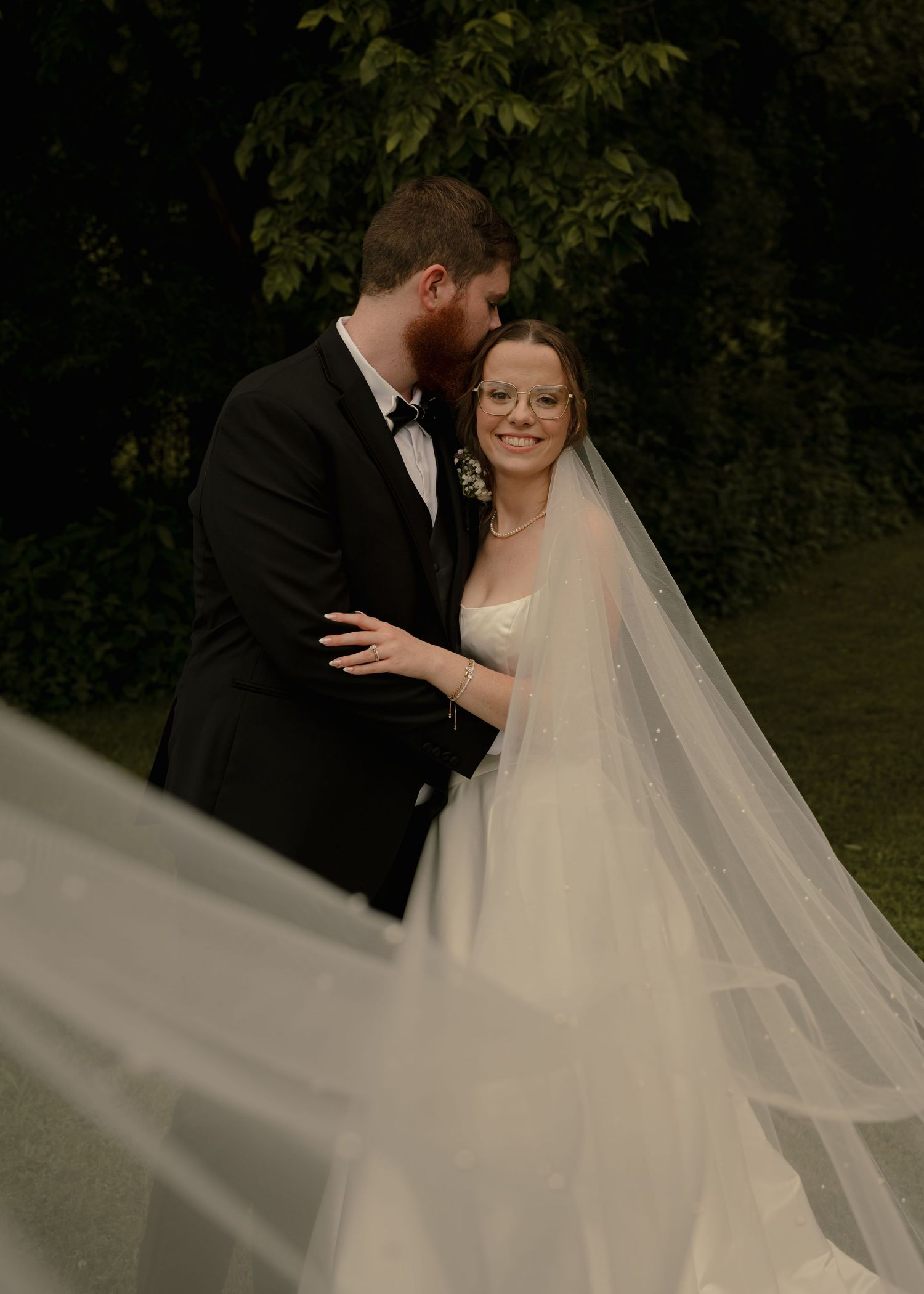 Groom in black tuxedo kissing bride wearing glasses and white veil in lush garden setting.