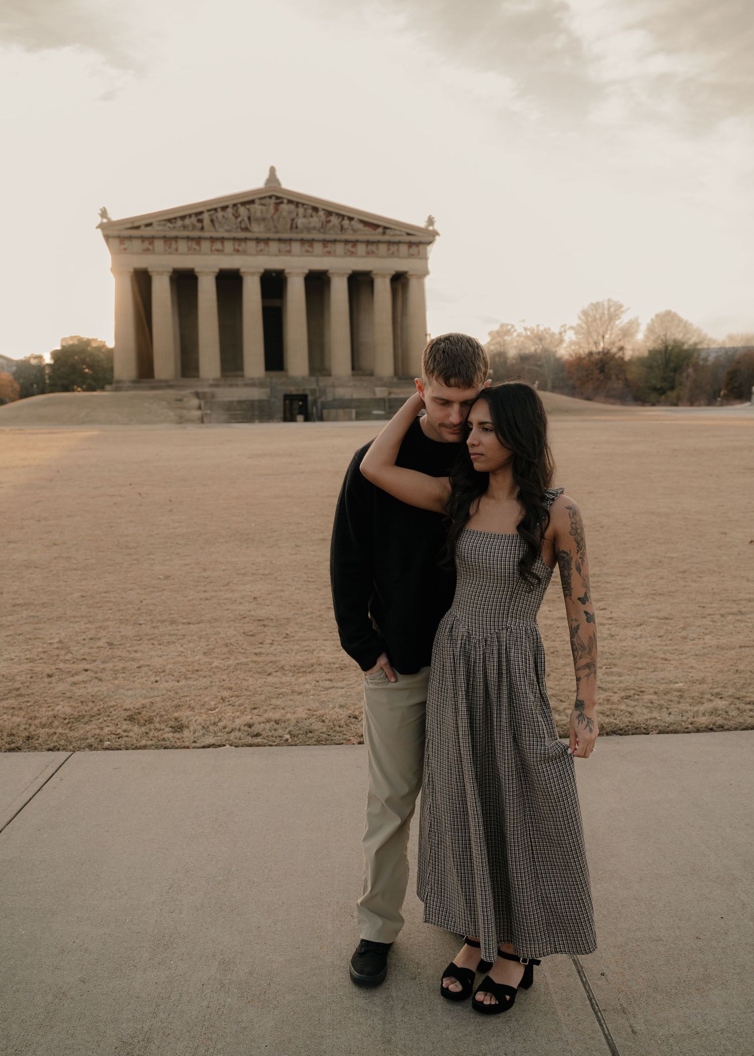 A couple poses together in front of the Nashville Parthenon, the woman in a gingham midi dress with visible arm tattoos.