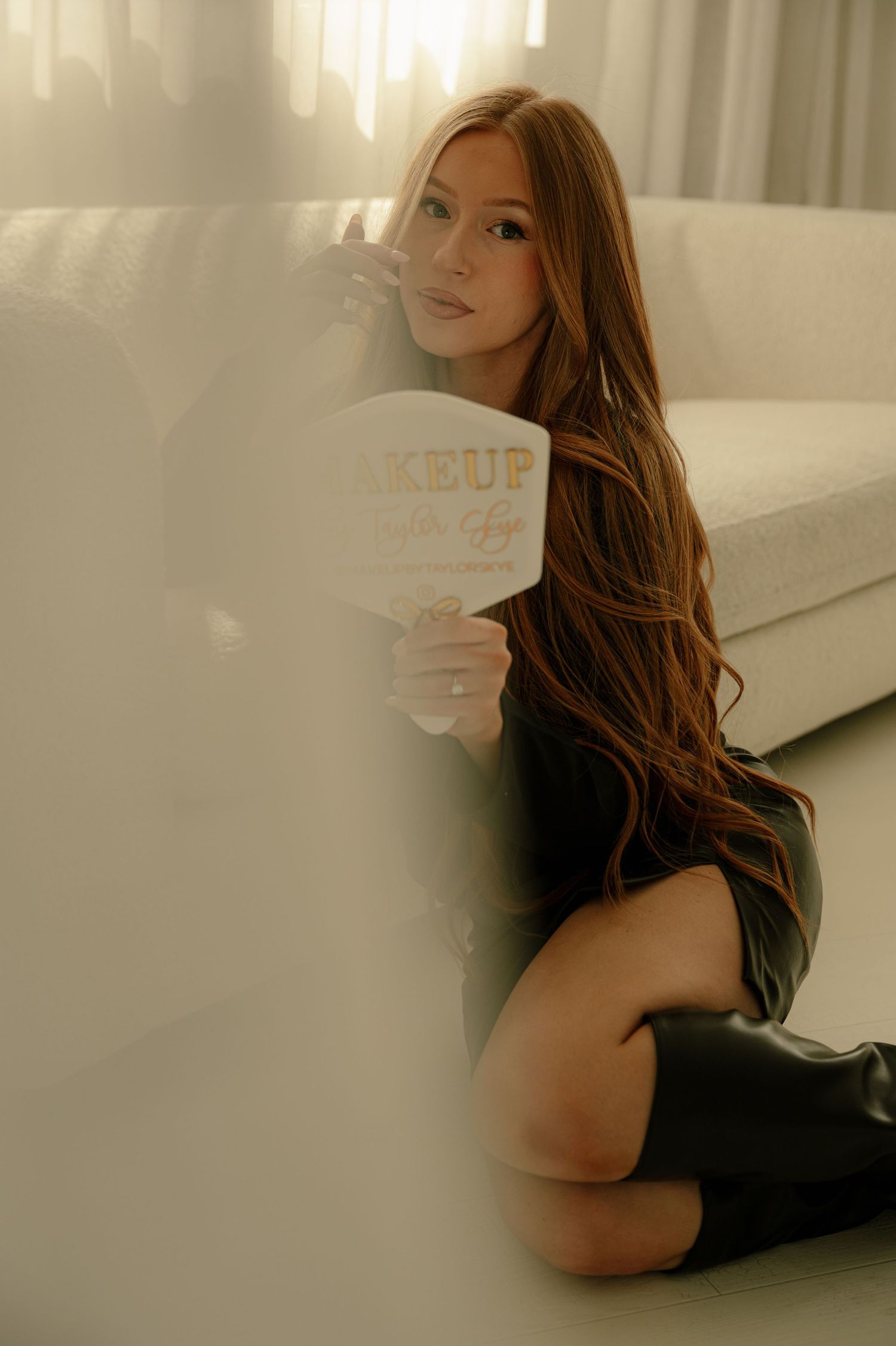 Woman with long auburn hair kneeling beside white sofa holding a makeup brand sign indoors.