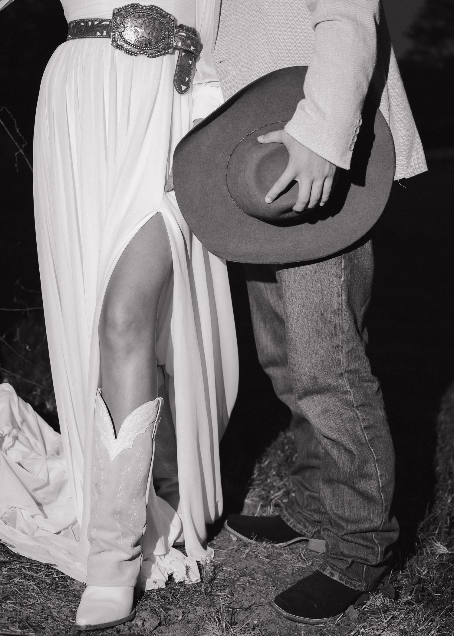 Bride in white dress with cowboy boots and belt beside groom holding cowboy hat, black and white.