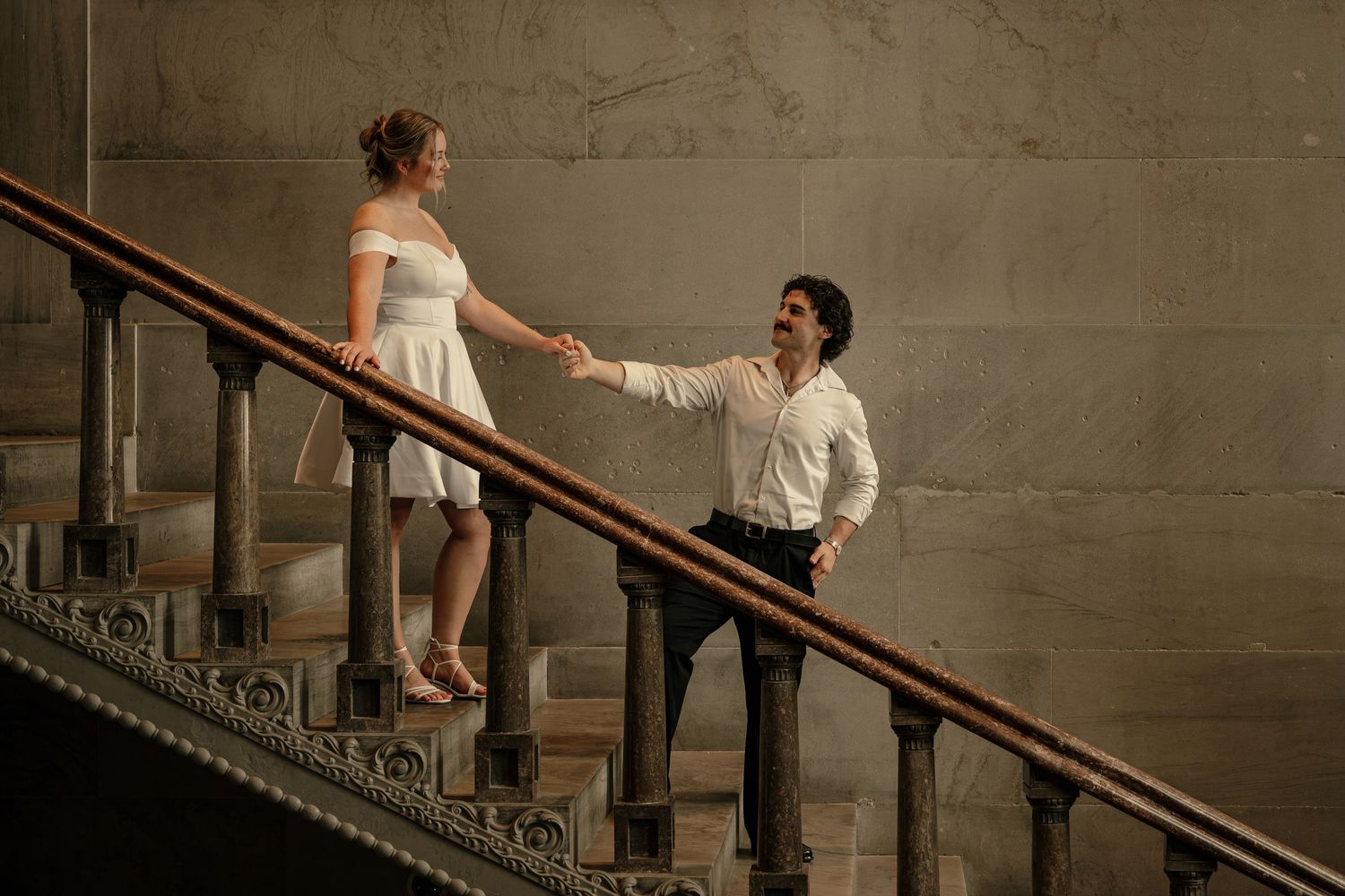 Couple holding hands on ornate staircase, woman in white dress and man in white shirt, elegant indoor setting.