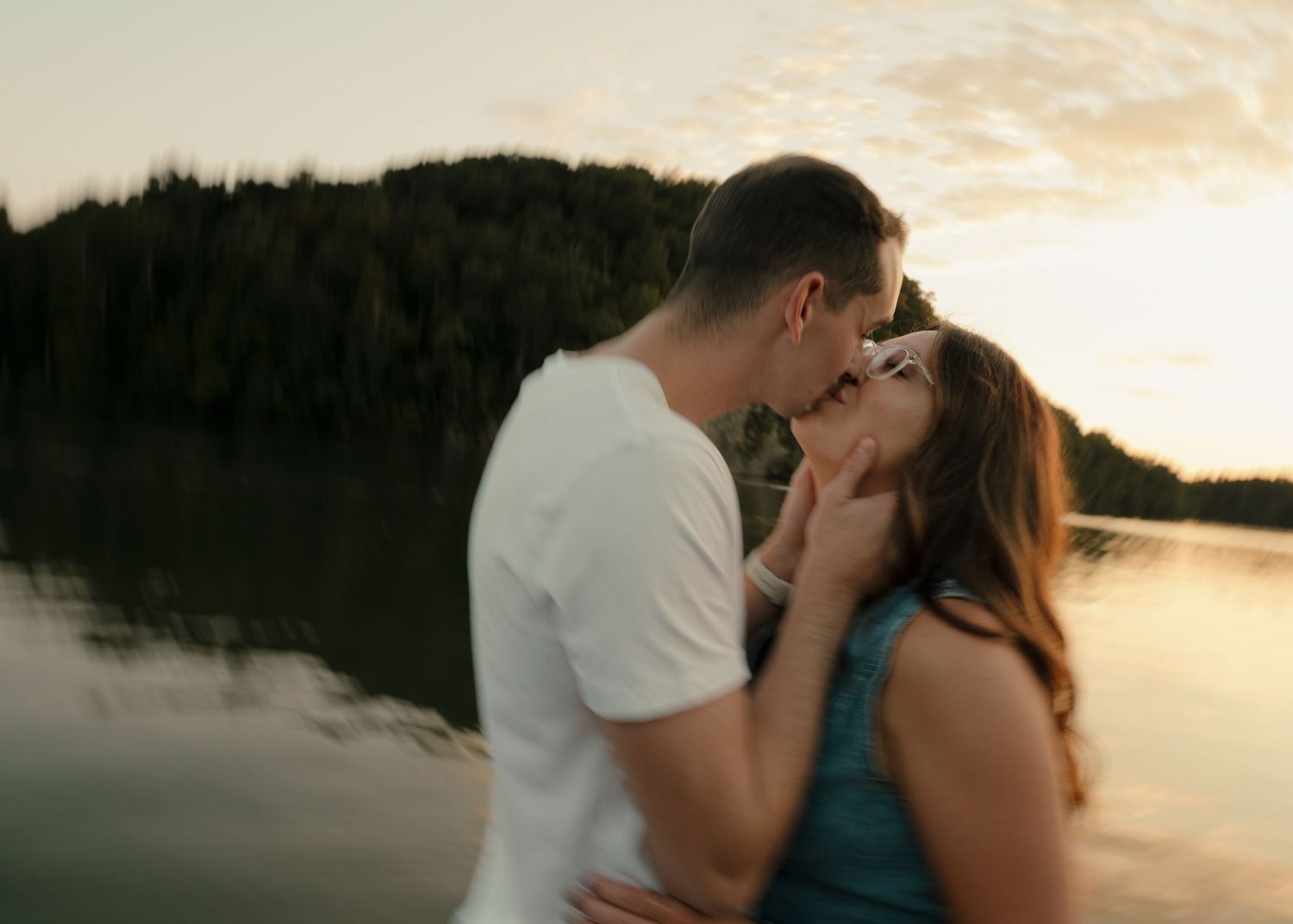 Couple kissing with a serene lake and glowing sunset sky behind them.