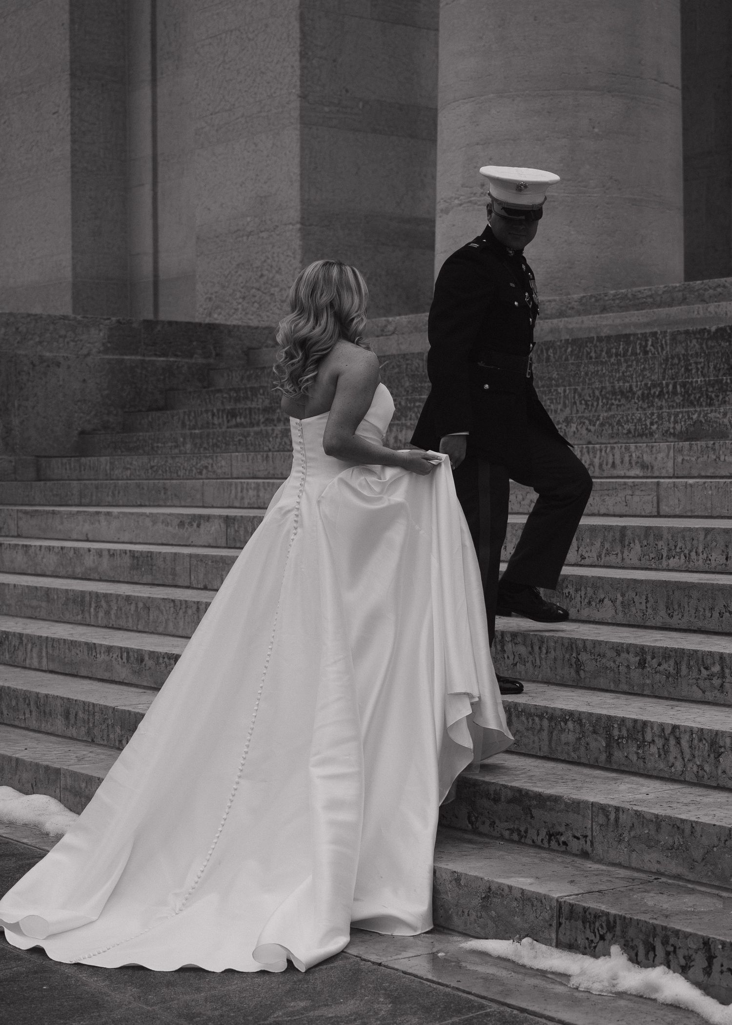 Black and white photo of bride in ball gown and military groom ascending stone steps at formal wedding.