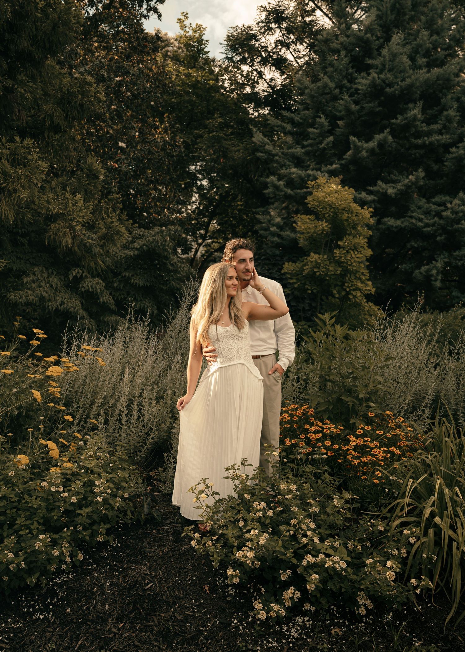 A couple embraces in a lush garden surrounded by wildflowers and greenery, the woman in a white dress.