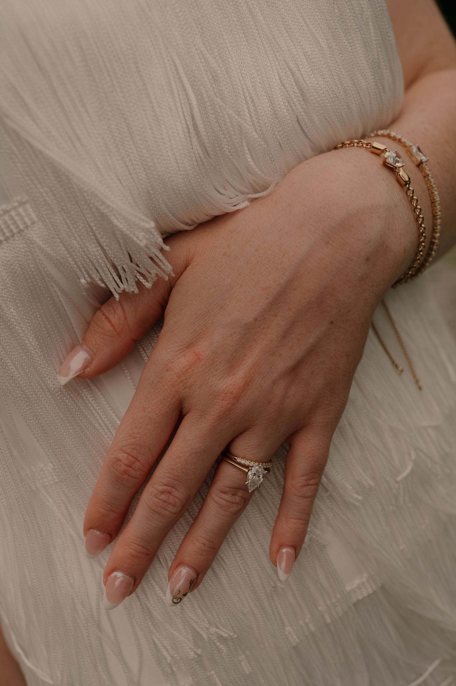 Close-up of bride's hand showing pear diamond ring and gold bracelet stack against white wedding dress.