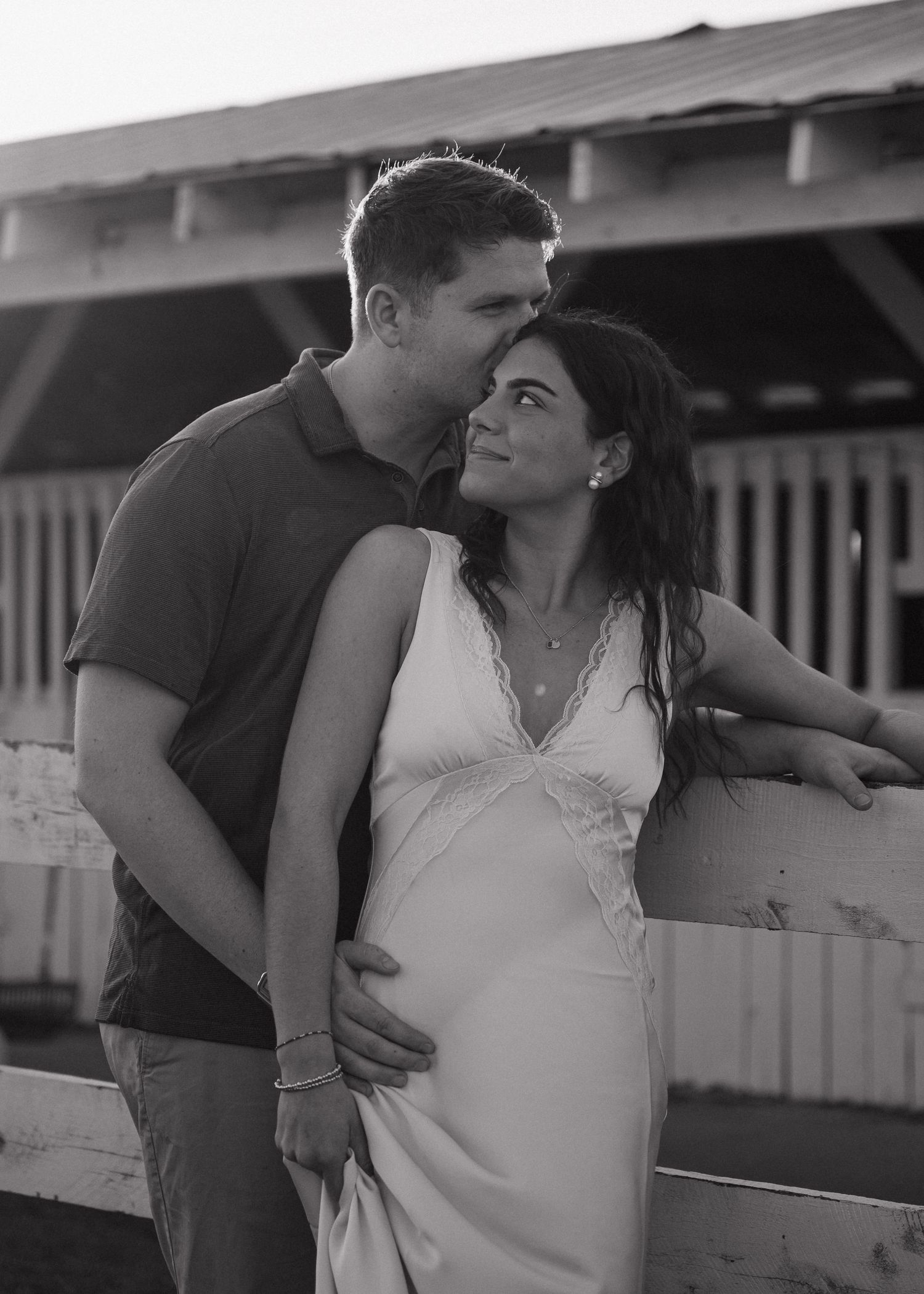 Black and white photo of couple embracing near white fence with woman in slip dress.