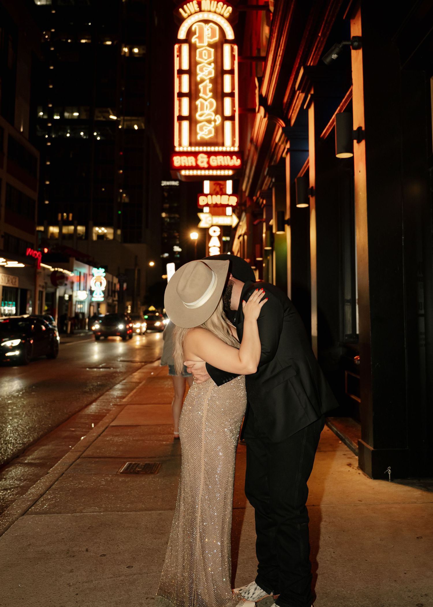 Couple kissing on a city sidewalk at night near a neon-lit bar and grill sign, dressed in elegant formal attire.