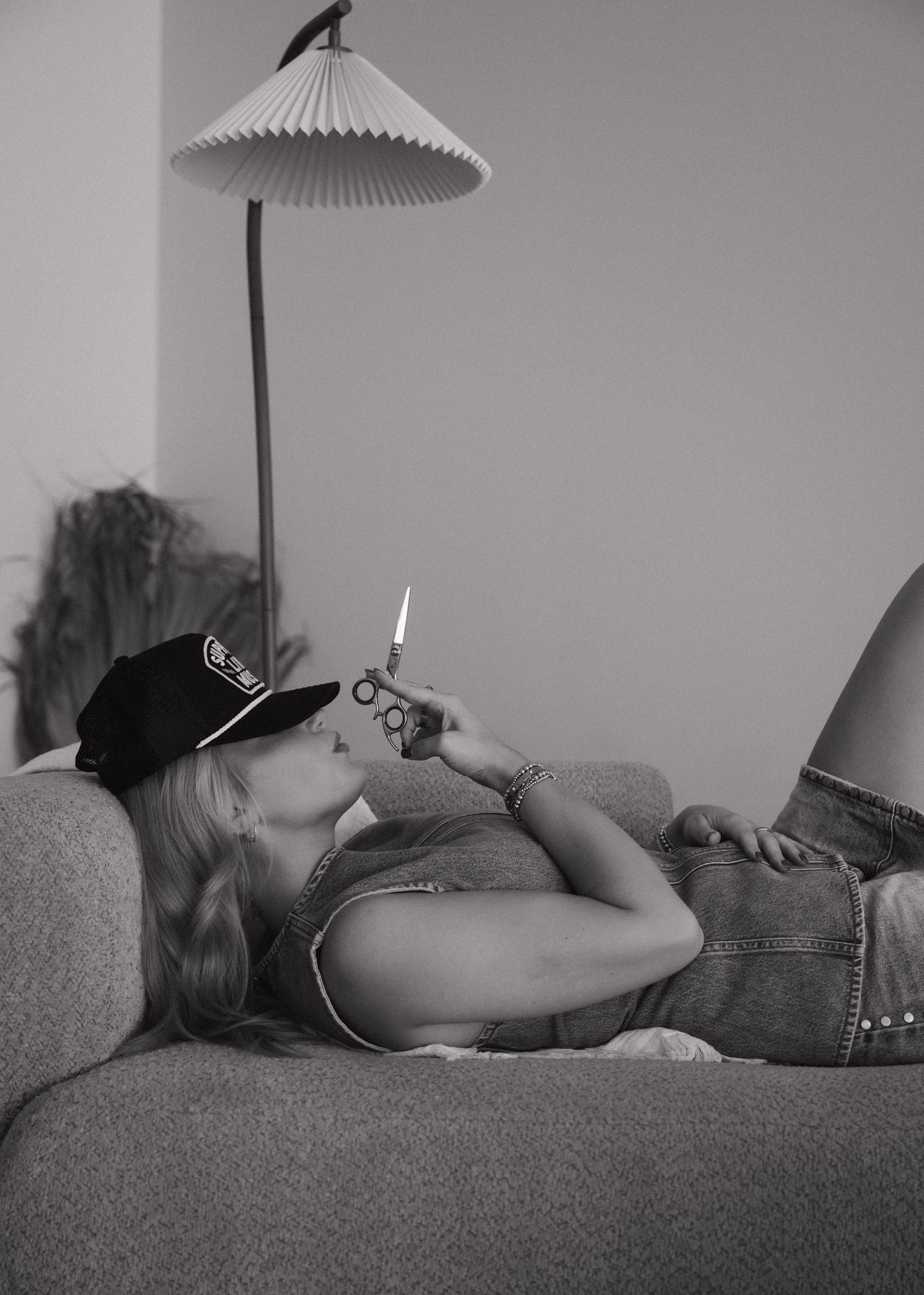 Black and white photo of a woman lying on a sofa, wearing a cap, holding a lighter in a retro-styled room.