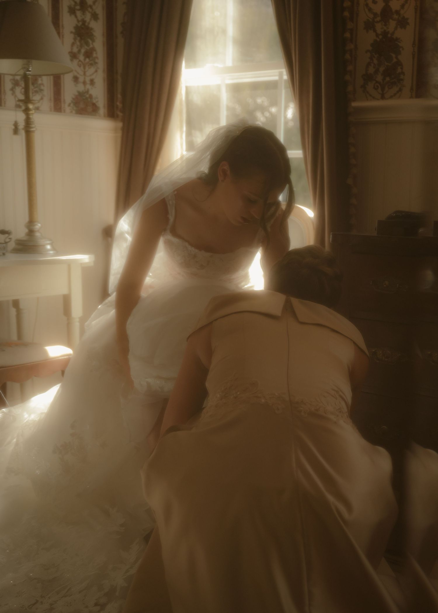 Bride in white ball gown leaning over seated person in sunlit vintage room with floral wallpaper.