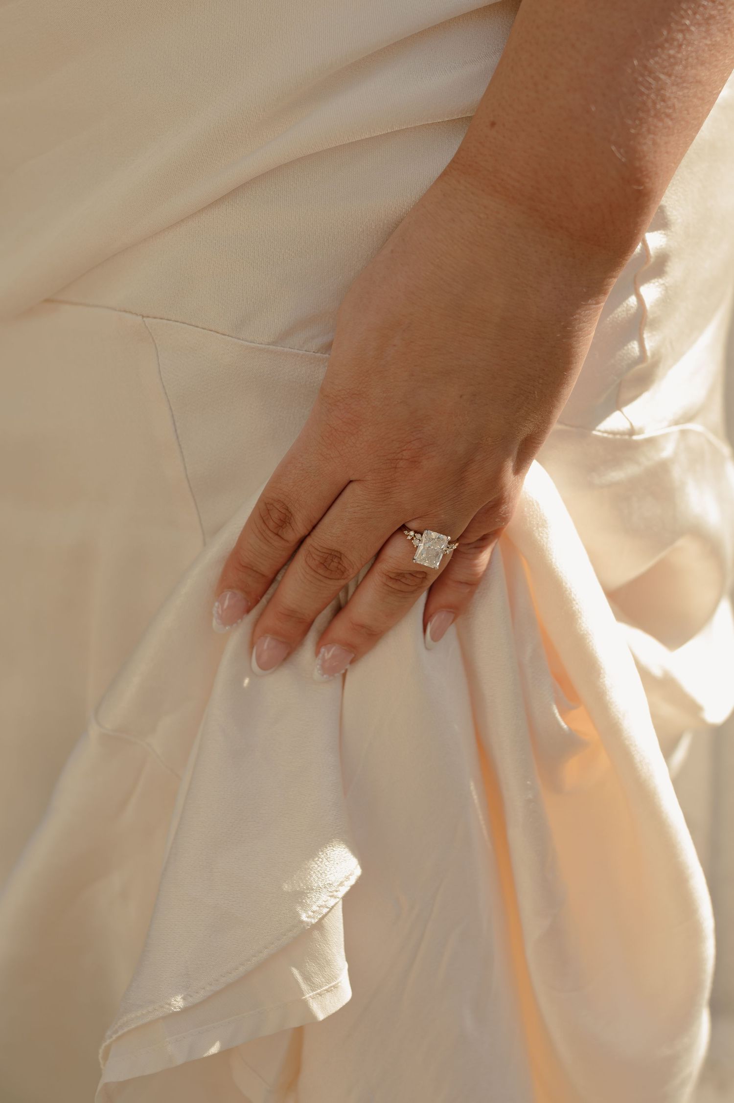 Close-up of woman's hand wearing a large emerald-cut diamond engagement ring on a cream dress.