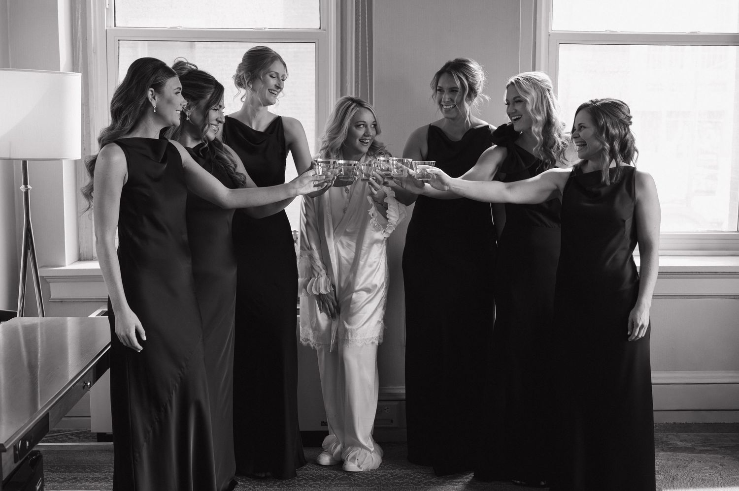 Black and white photo of bride in robe toasting with bridesmaids in black dresses before wedding.