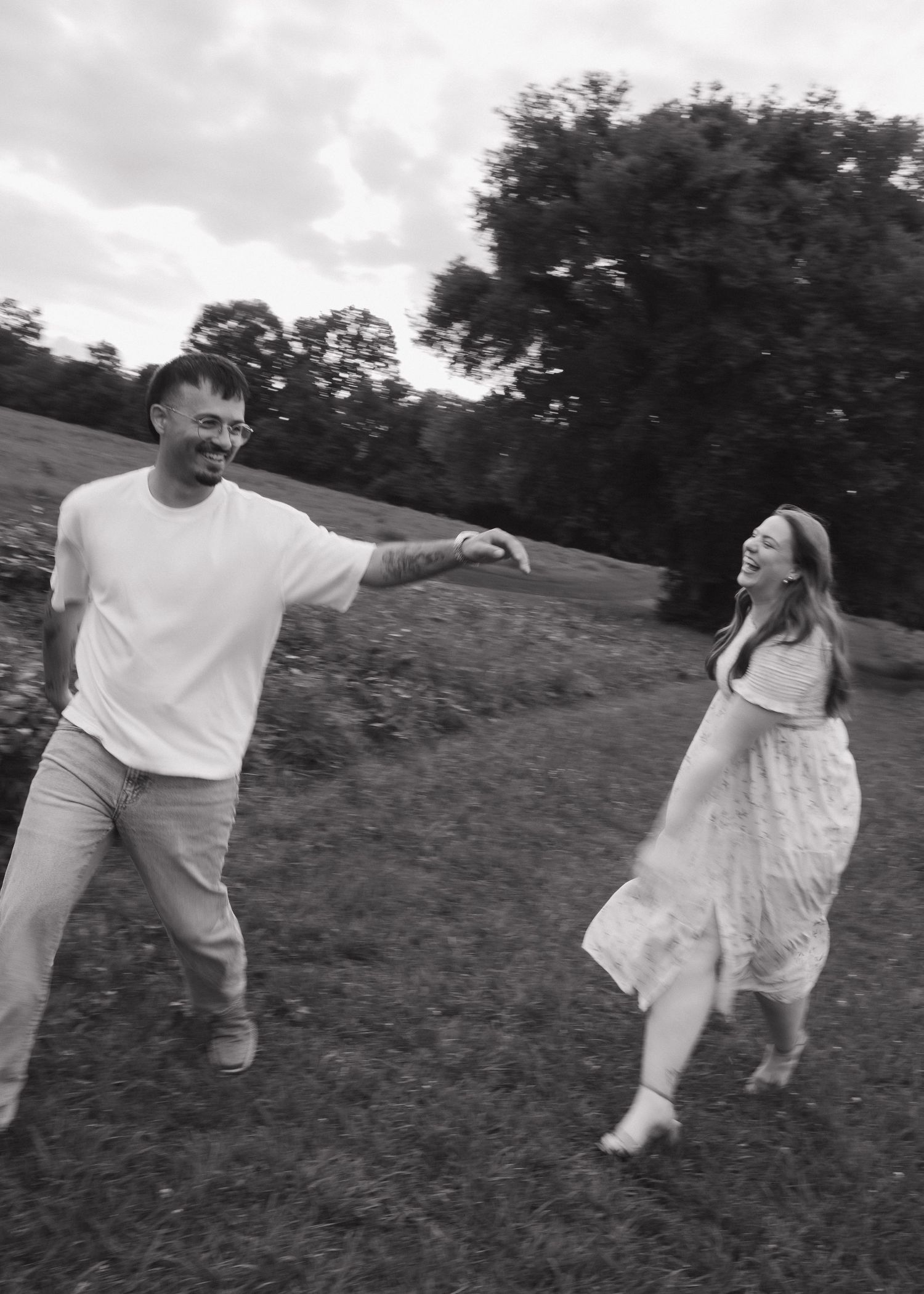 Black and white photo of a joyful couple dancing and laughing together in a lush green outdoor field.