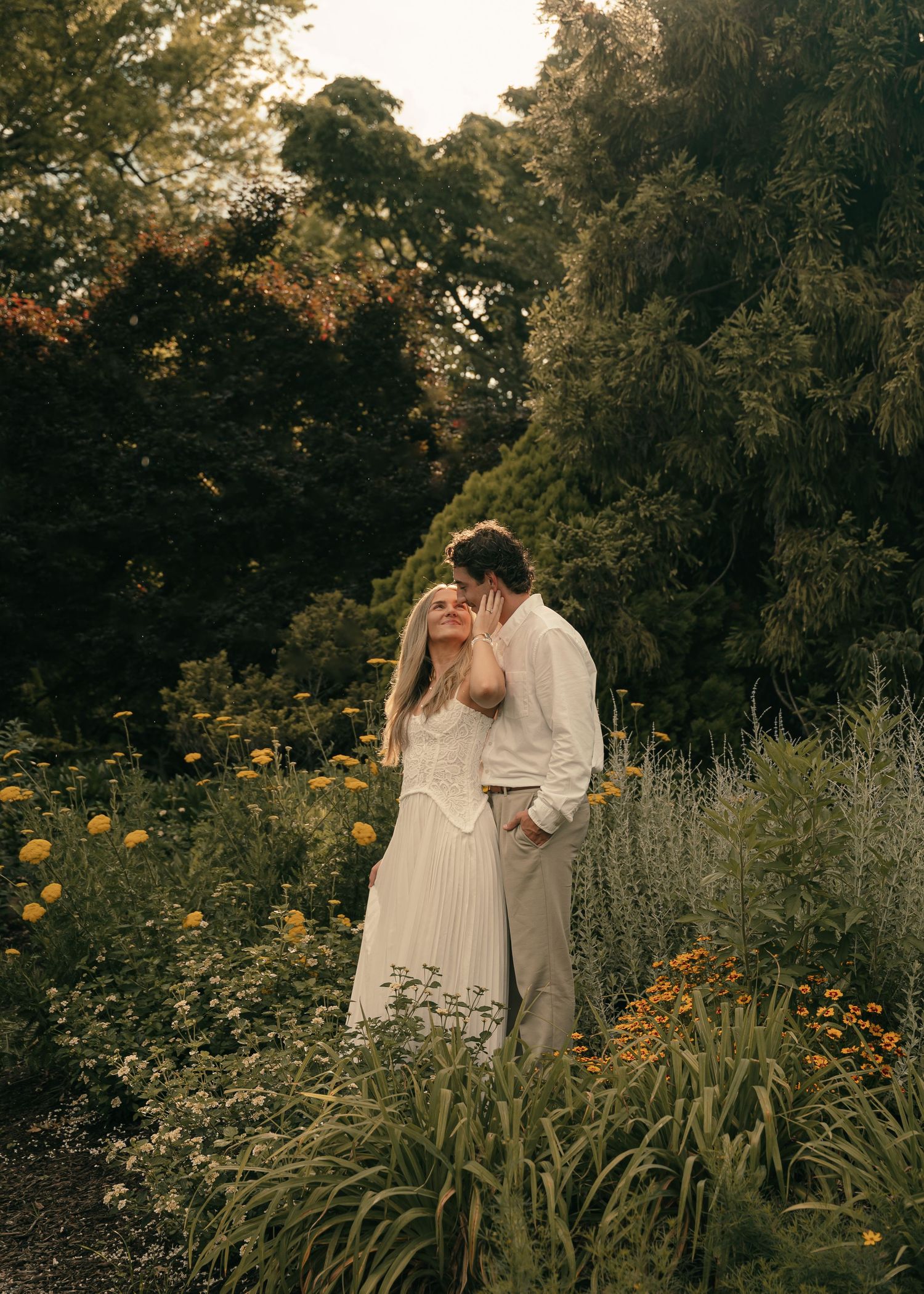 Couple embracing in a lush garden surrounded by wildflowers and greenery during golden hour.