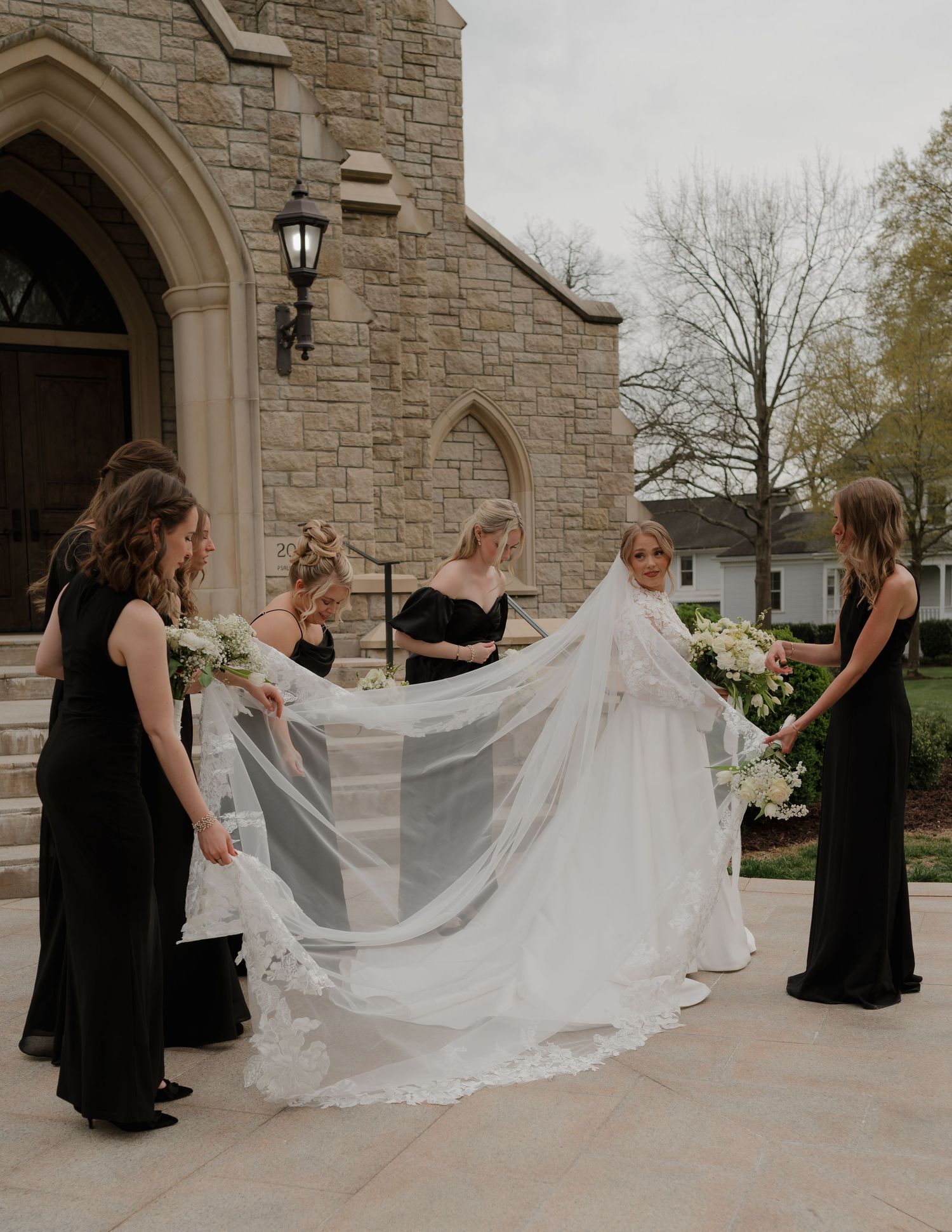 Bride in lace gown with bridesmaids in black dresses holding veil outside stone church.