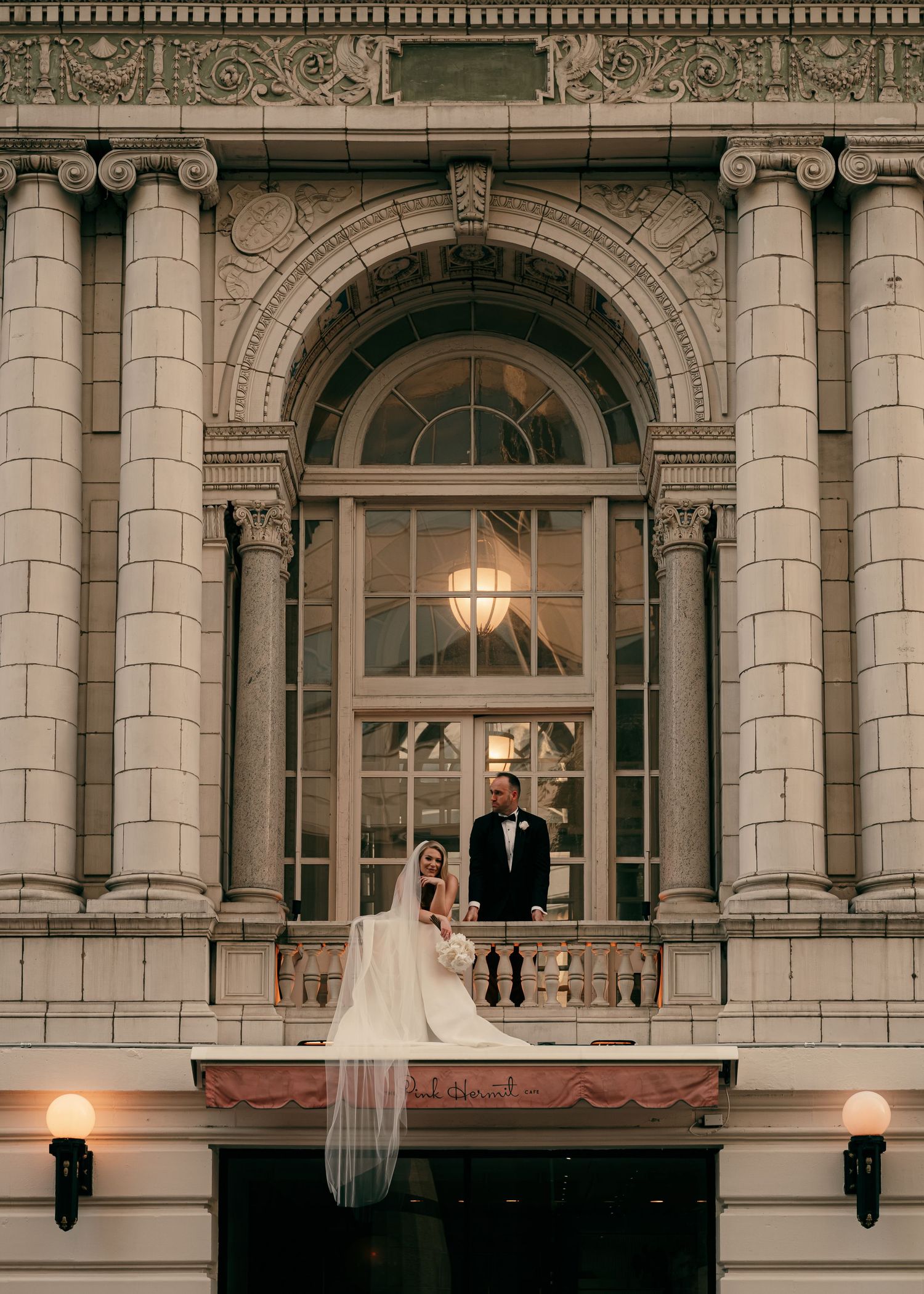 Bride and groom stand on ornate building balcony, she in white gown with veil, he in black tux, at elegant wedding venue.