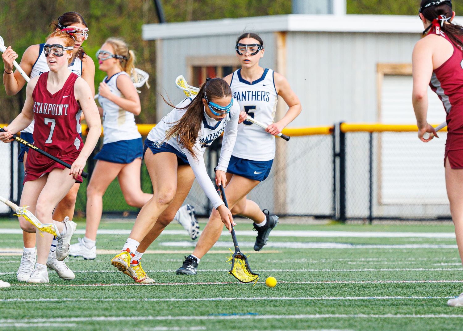 Girls lacrosse players in white Panthers uniforms compete on a turf field during a game against a team in red uniforms.