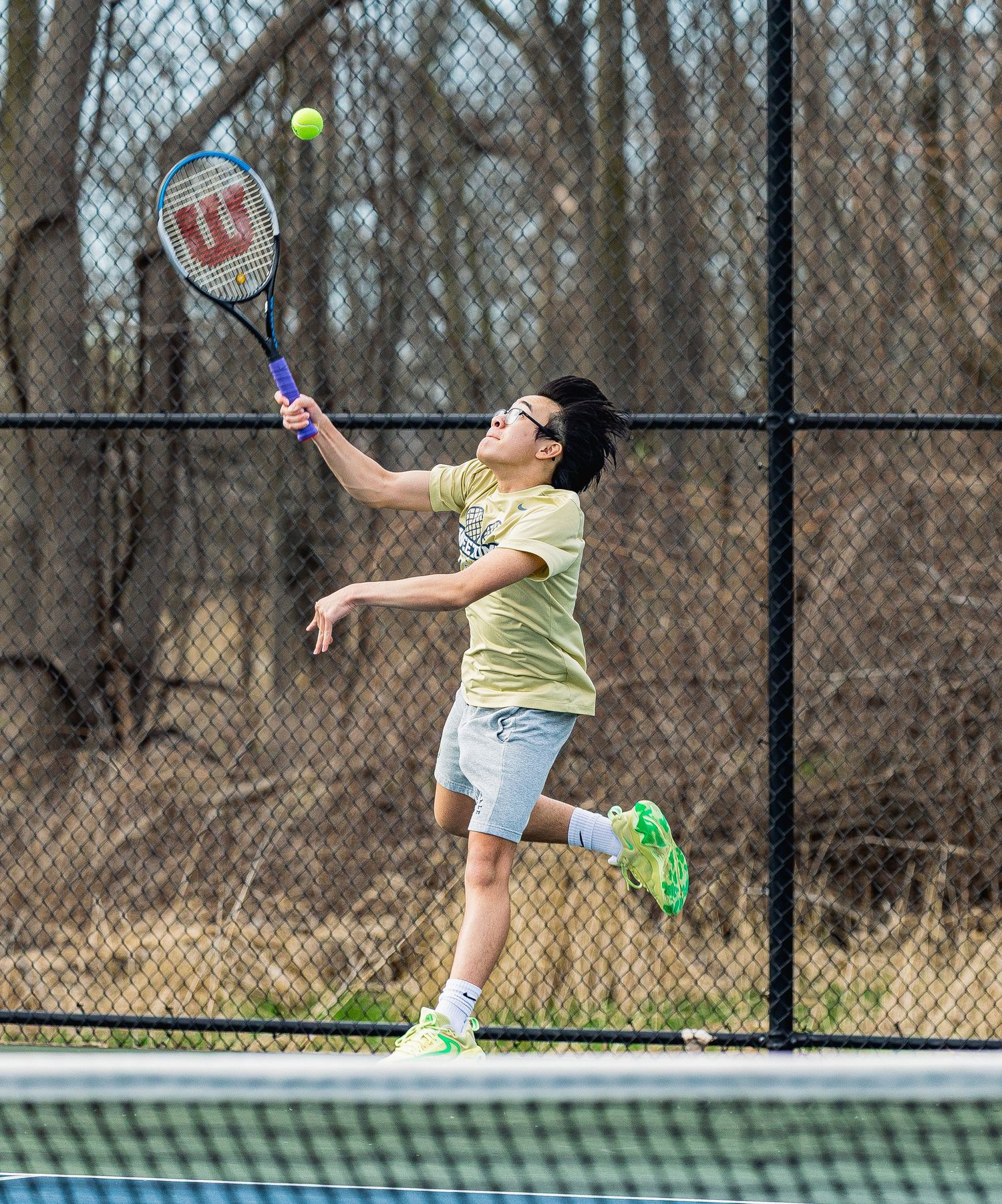Young tennis player hitting an overhead shot with a Wilson racket on an outdoor court.