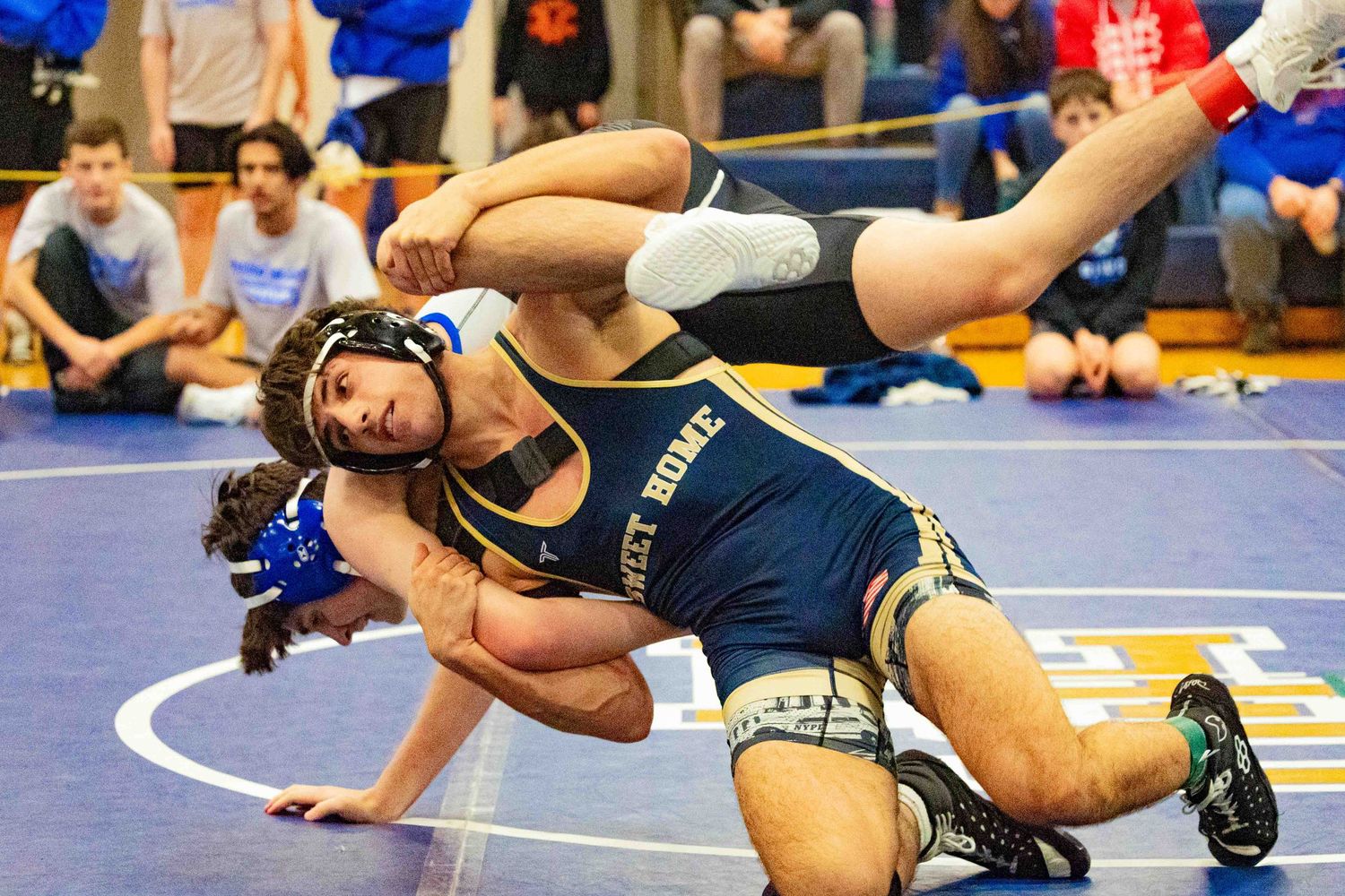 Two wrestlers compete on a blue mat, one in a navy Jersey Shore singlet executing a takedown move.