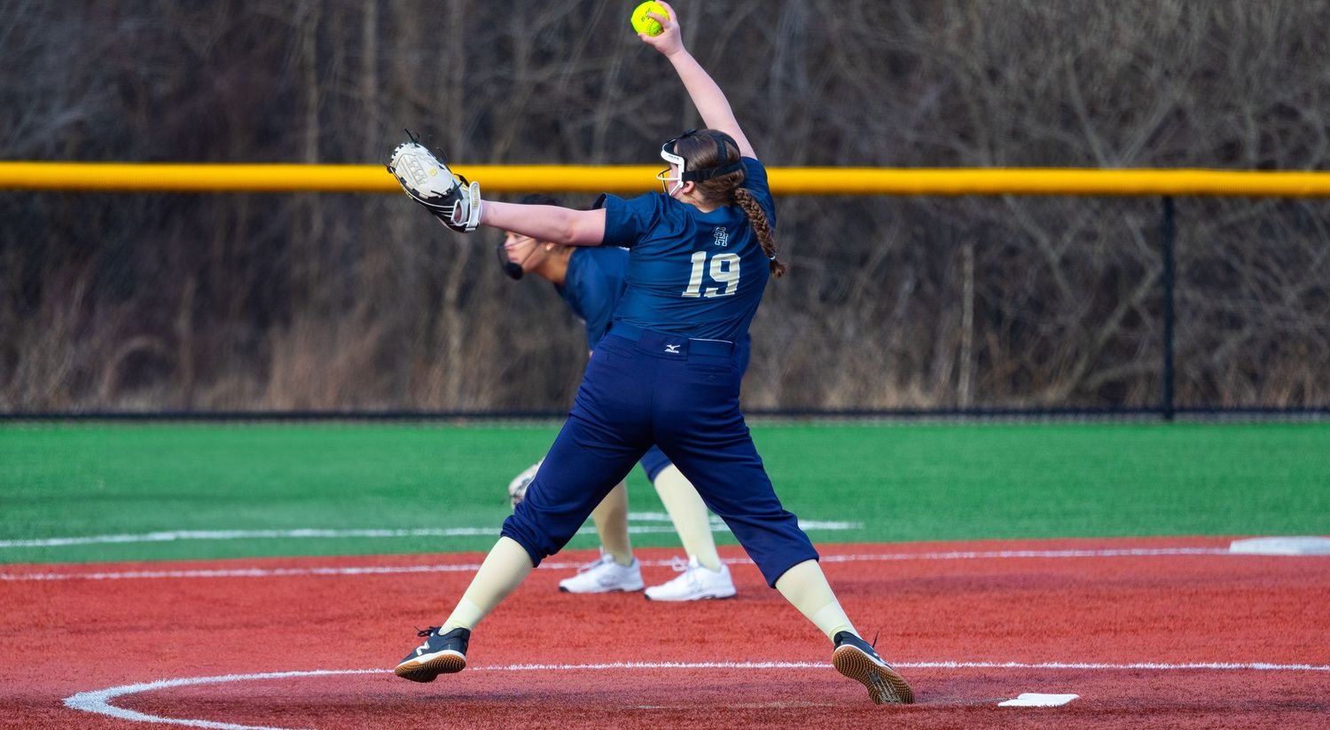 Softball pitcher in navy uniform mid-delivery on pitcher's mound.