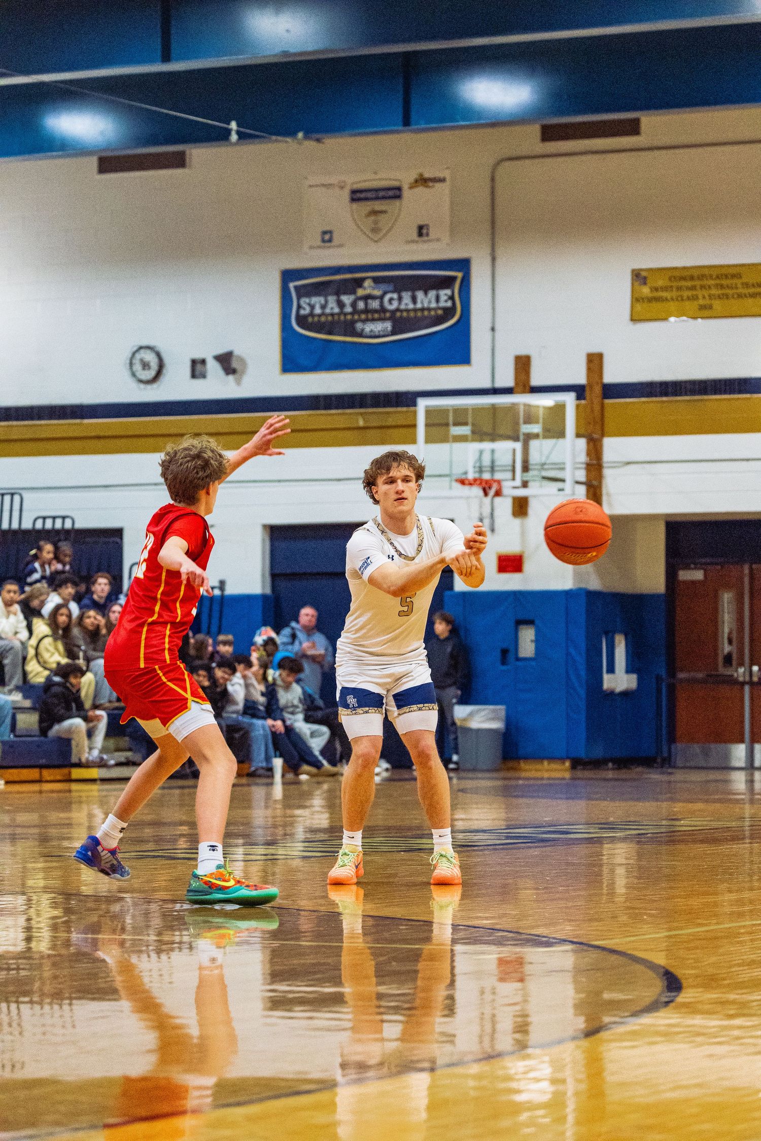 High school basketball player #5 in blue uniform passes ball during indoor game against red-uniformed opponent.