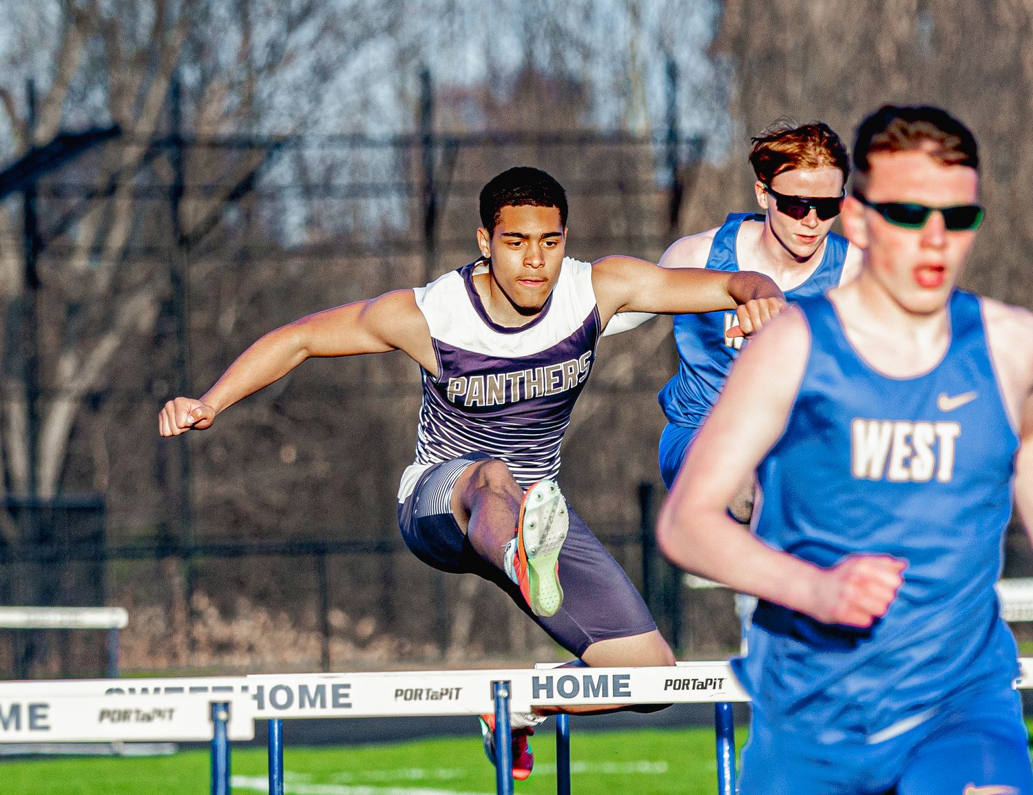 A Panthers male athlete leaps over a hurdle ahead of competitors during an outdoor track meet.