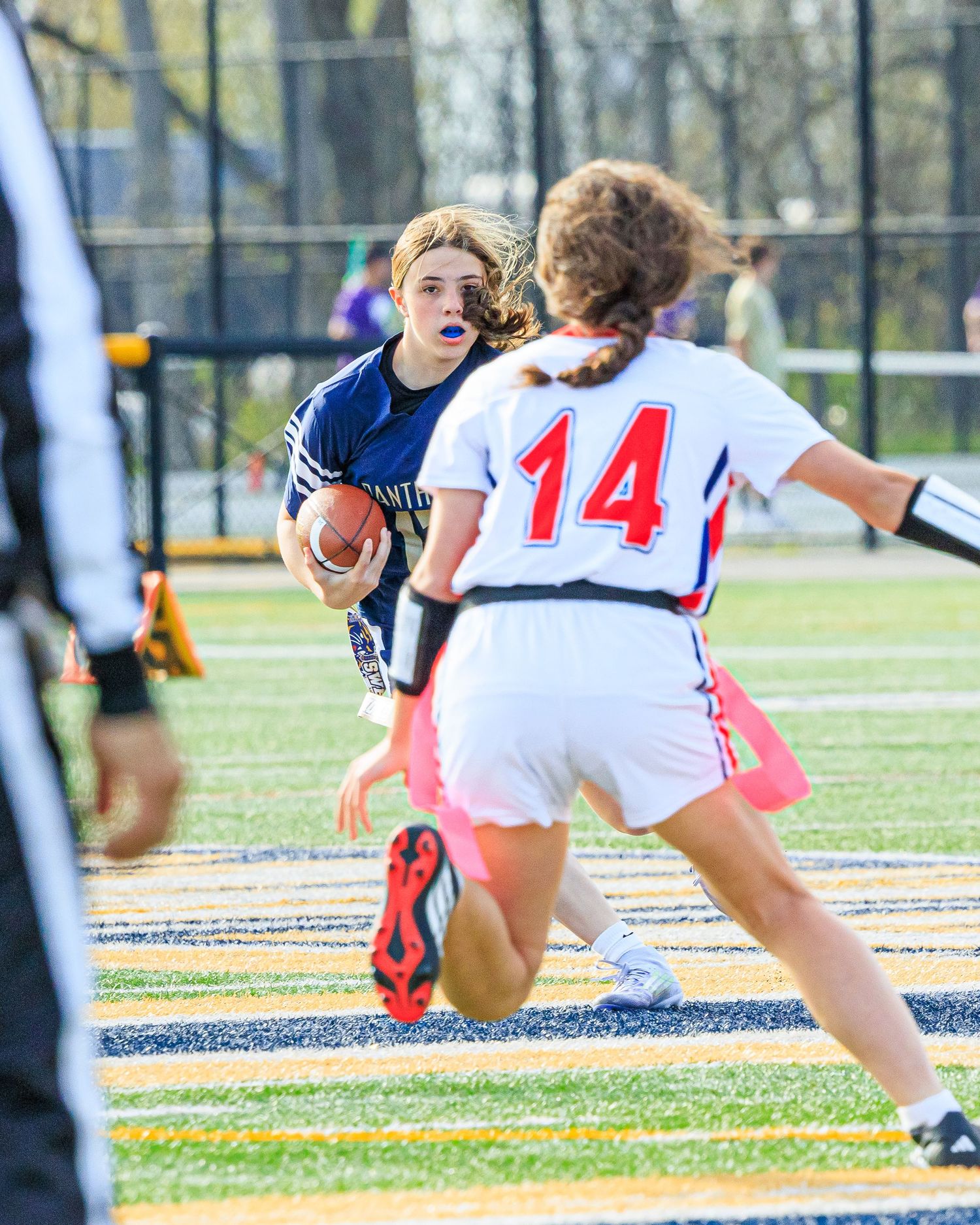 Girl in navy uniform runs with football while opponent in number 14 jersey defends.
