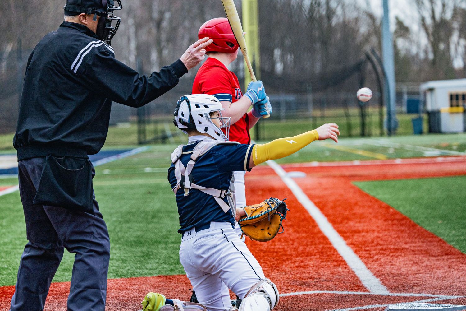 Baseball catcher in navy uniform catches pitch as red-uniformed batter swings near home plate.