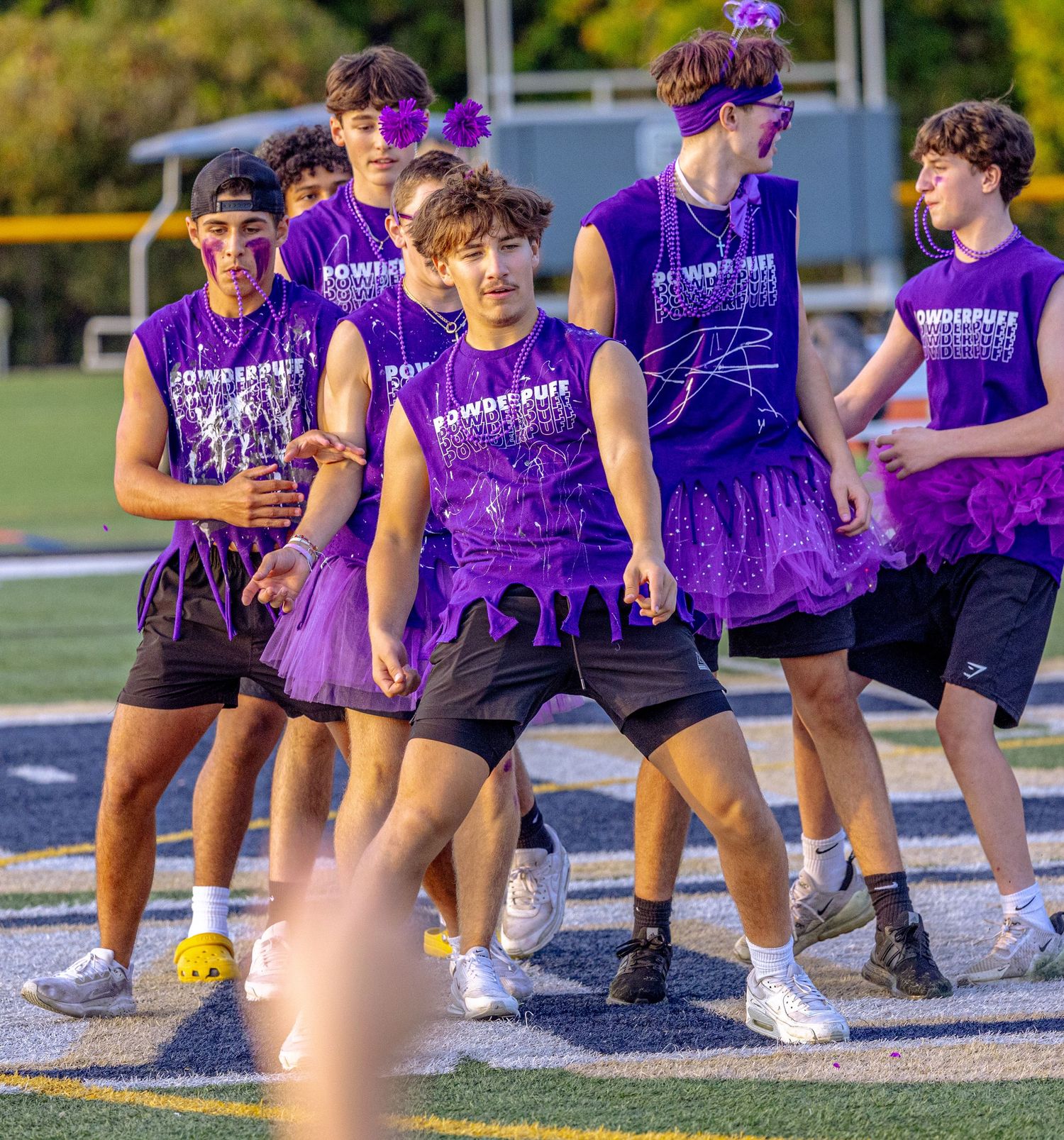 Teen boys in purple Powderpuff shirts dance on a football field during a school spirit event.