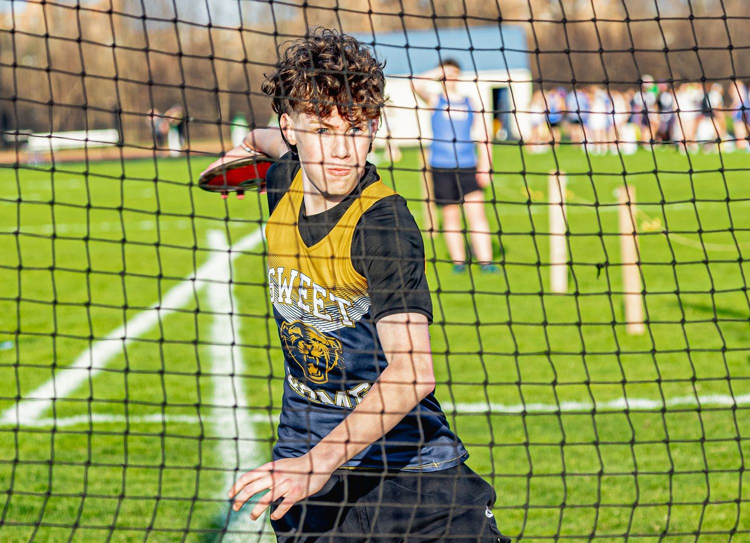 Young athlete in yellow vest throws discus on green turf field during outdoor track and field competition.