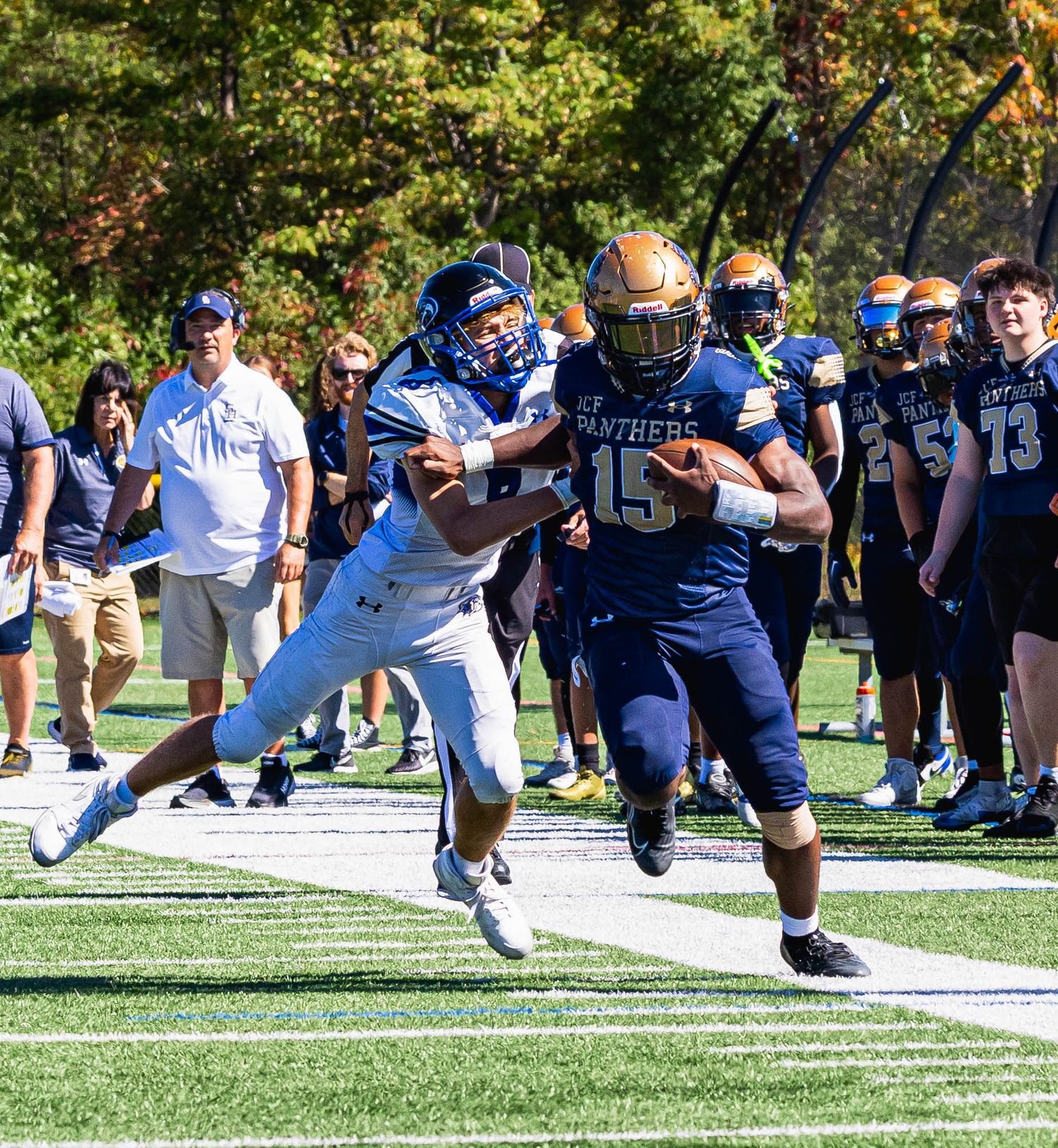 Football players collide on a sunny field during an intense play with spectators watching from the sidelines.