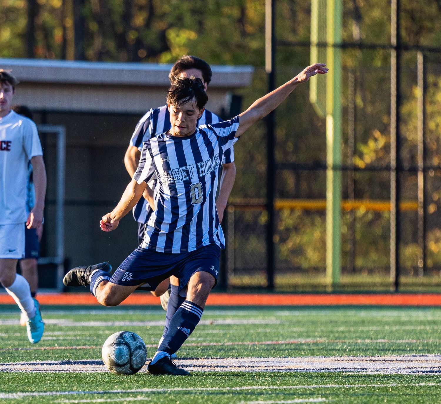 A soccer player in a striped jersey kicks the ball on a green turf field during a competitive match.