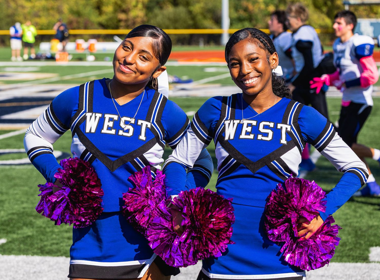 Cheerleaders in blue and white uniforms perform with purple pom poms during an outdoor sporting event.