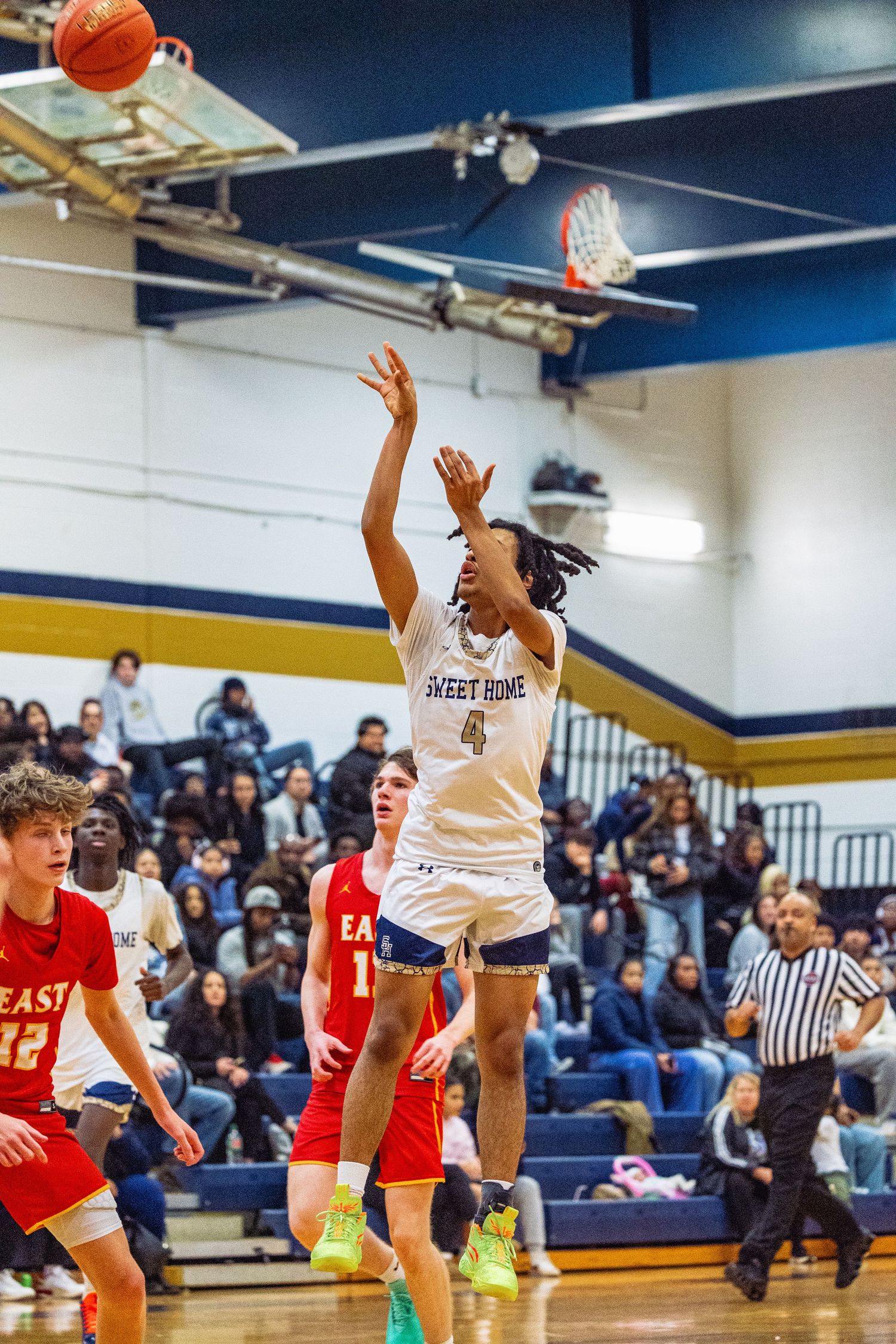 West Home basketball player #4 in white uniform jumps to shoot ball during game against East team in red uniforms.