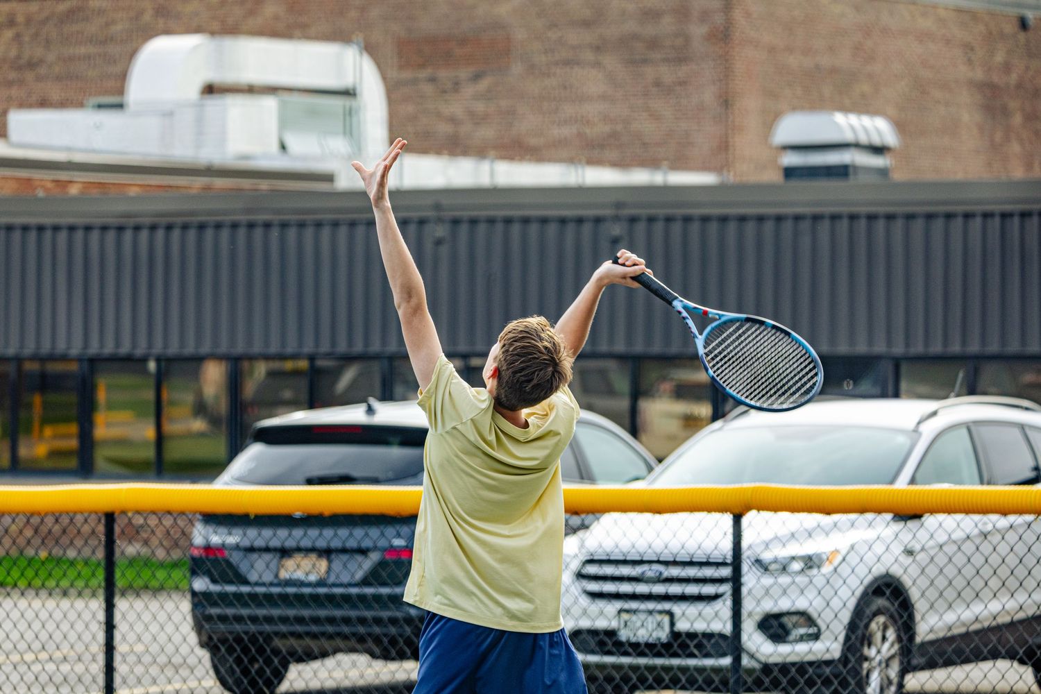 A tennis player raises their racket overhead on an outdoor court near a parking lot with cars and a brick building.