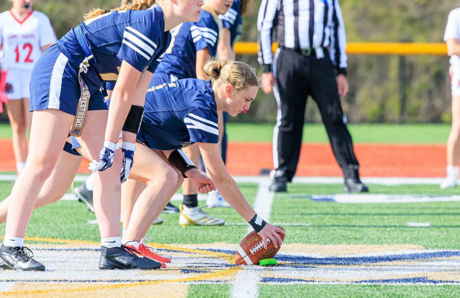 Young female athlete snaps football during a flag football game on a turf field wearing blue uniforms.
