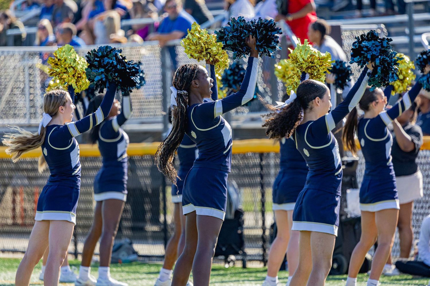 Cheerleaders in navy blue uniforms perform with yellow pom-poms during an outdoor sporting event.