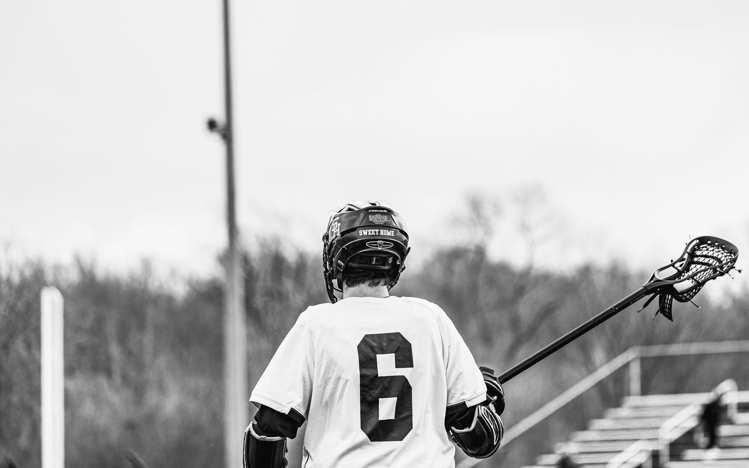 Black and white photo of lacrosse player number 6 holding a stick, viewed from behind on an outdoor field.