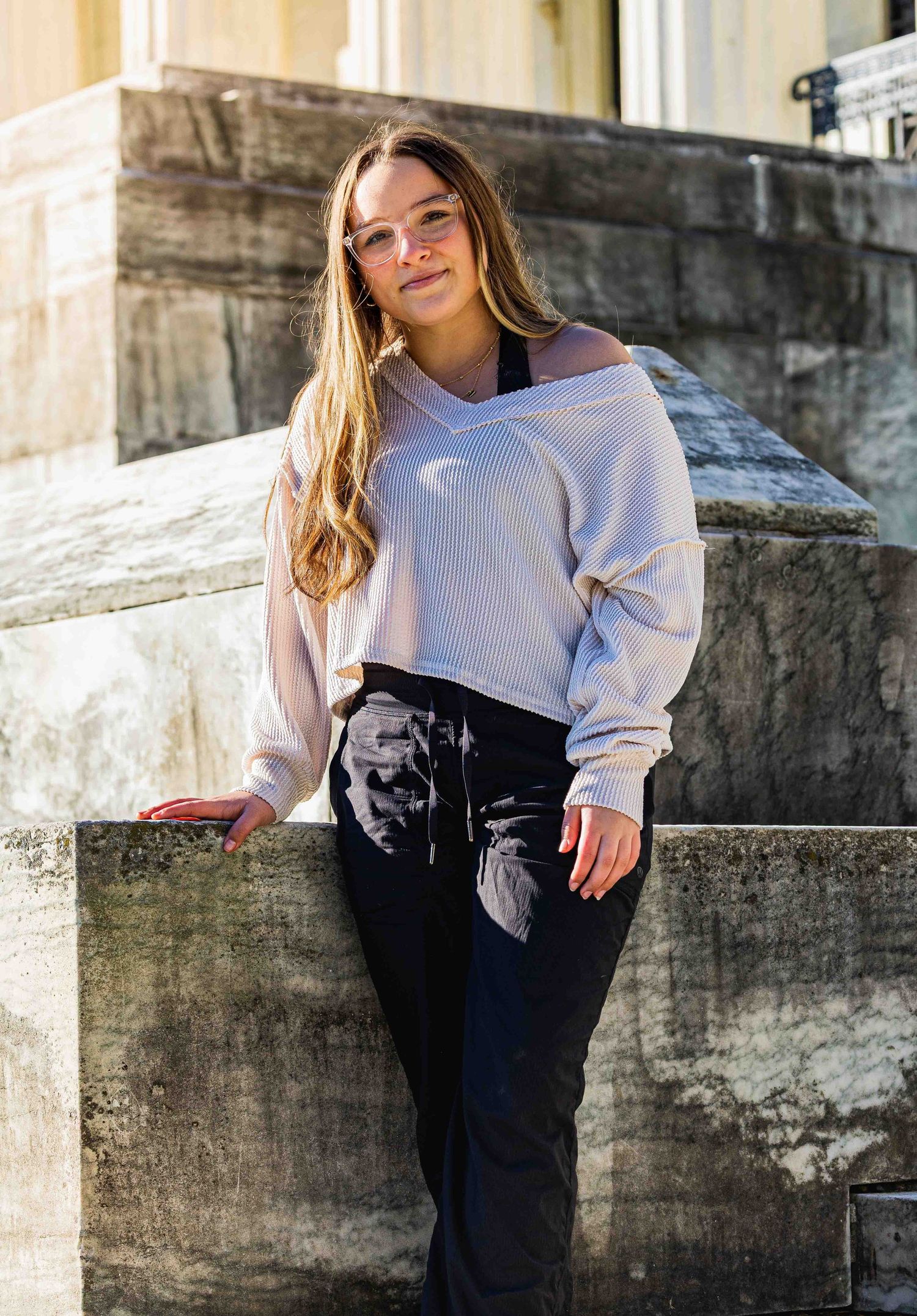 Young woman in white sweater and black floral pants leaning against stone wall outdoors.