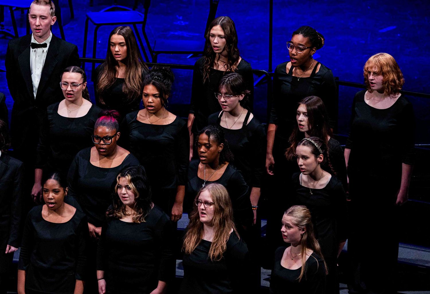 A large choir of young women in black dresses performing on stage under blue stage lighting.