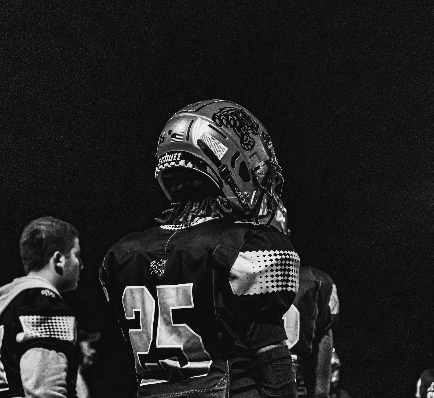 Black and white photo of football player number 25 wearing a helmet standing on the sideline during a game.