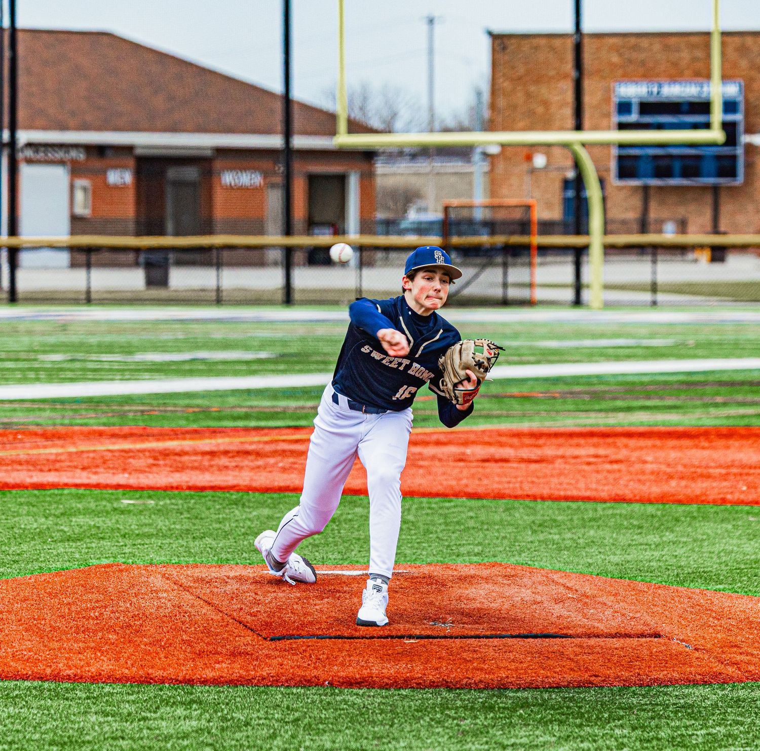 Young baseball pitcher in navy uniform delivering a pitch on an orange turf field during a game.