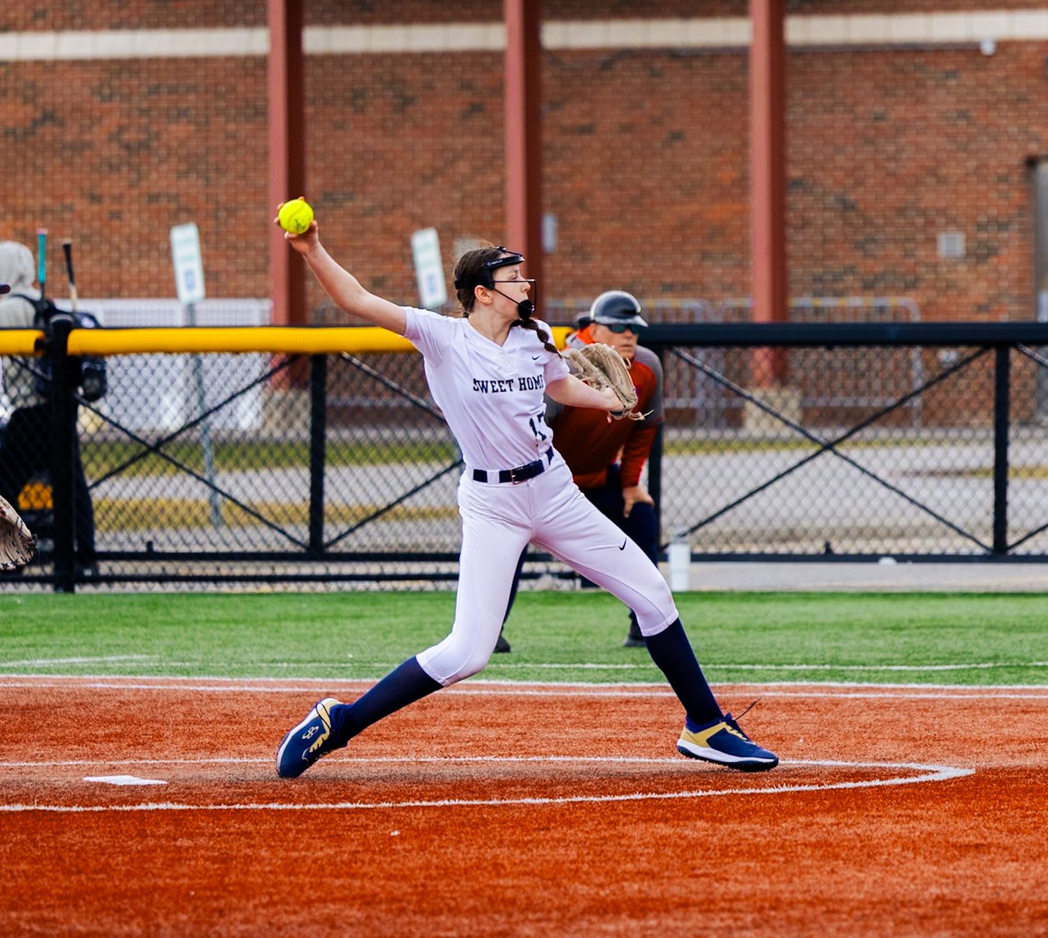 Softball player in white uniform stands at bat ready to hit yellow ball on dirt field with brick wall background.