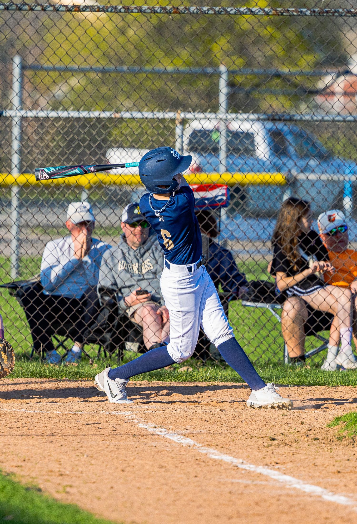 Baseball batter in navy uniform swings at pitch while spectators watch from behind chain link fence during daytime game.