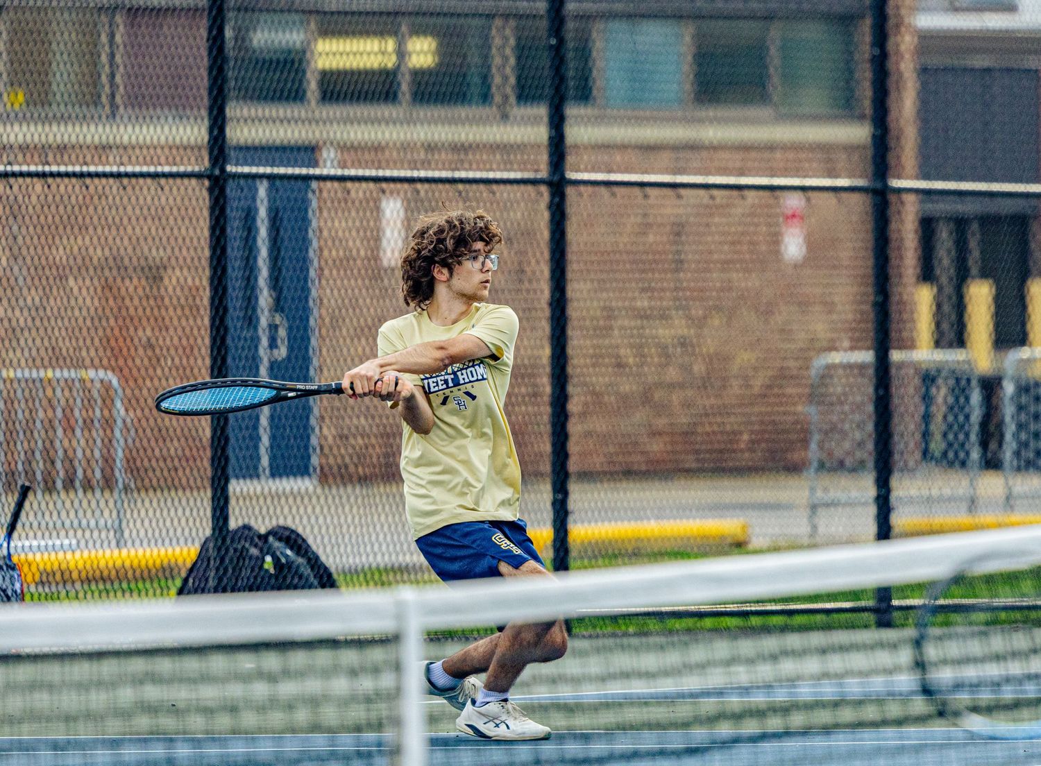 Young tennis player in yellow shirt hits a backhand shot on an outdoor court with chain-link fence backdrop.