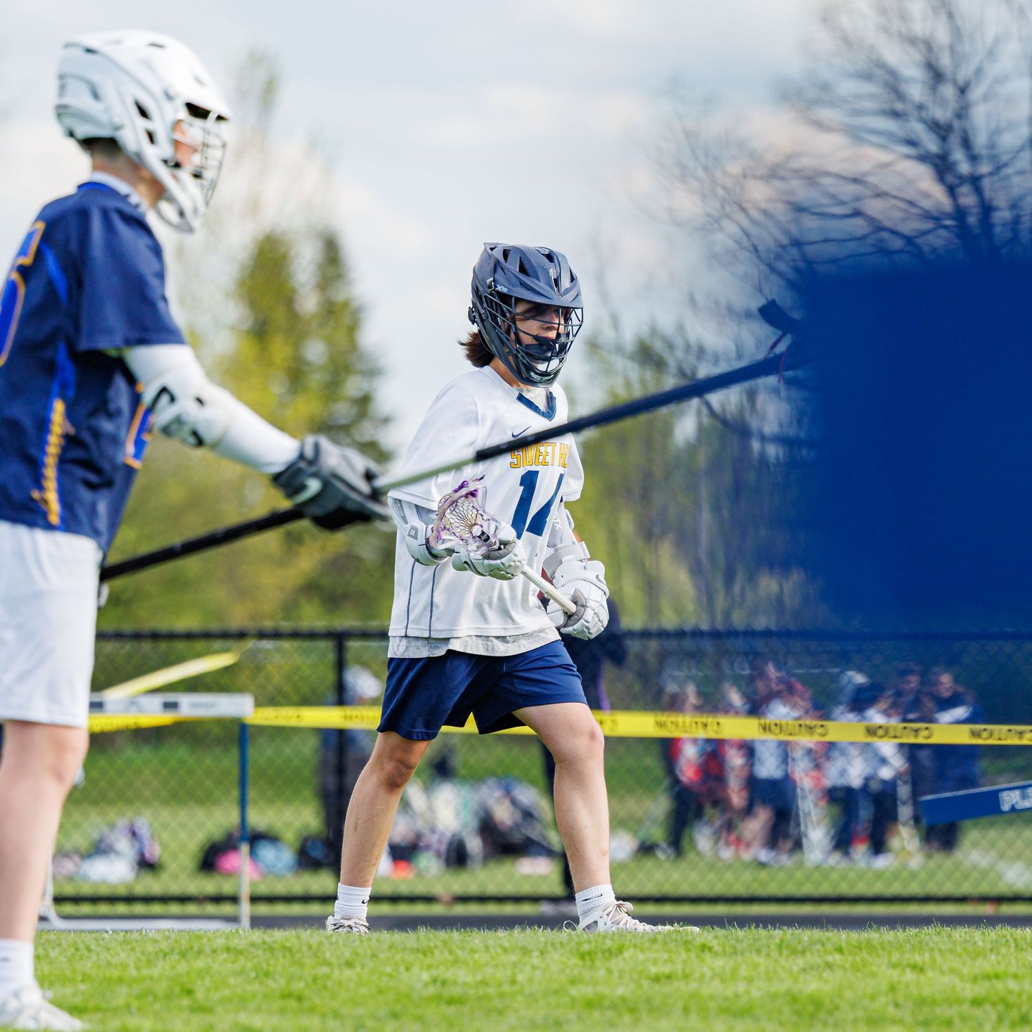Two youth lacrosse players face off on a green field during a spring game, wearing helmets and full protective gear.